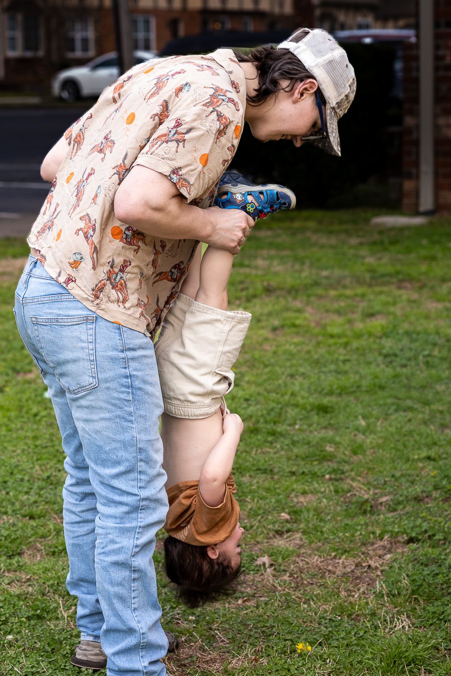 Father playfully holding toddler upside down by ankles on green grass outdoors.