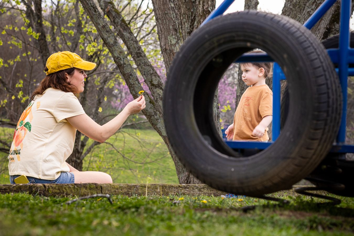 Mother in yellow cap photographing young boy seen through a tire swing in a park.