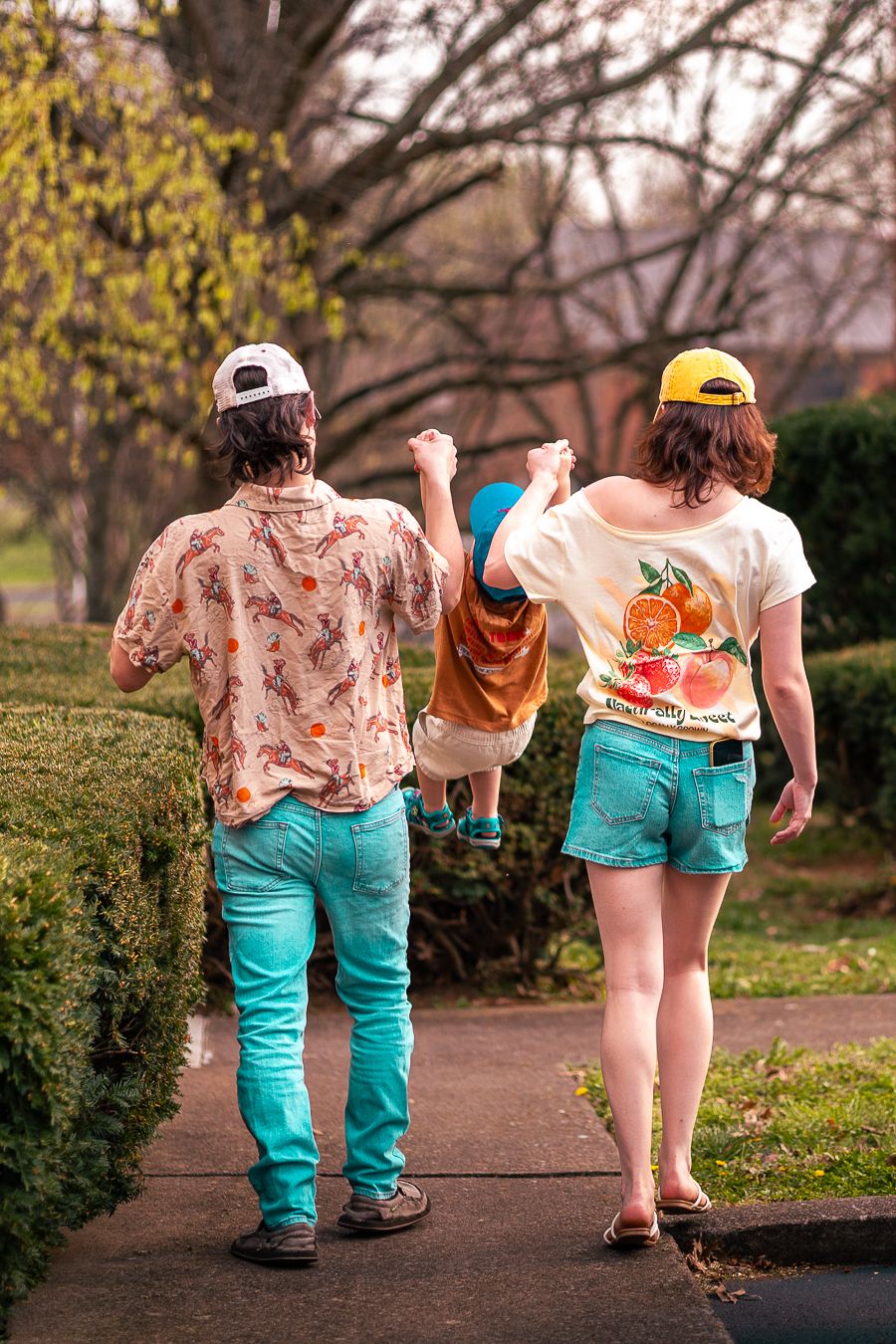Young family walking on sidewalk, parents holding toddler's hands between them, wearing matching caps and denim shorts.