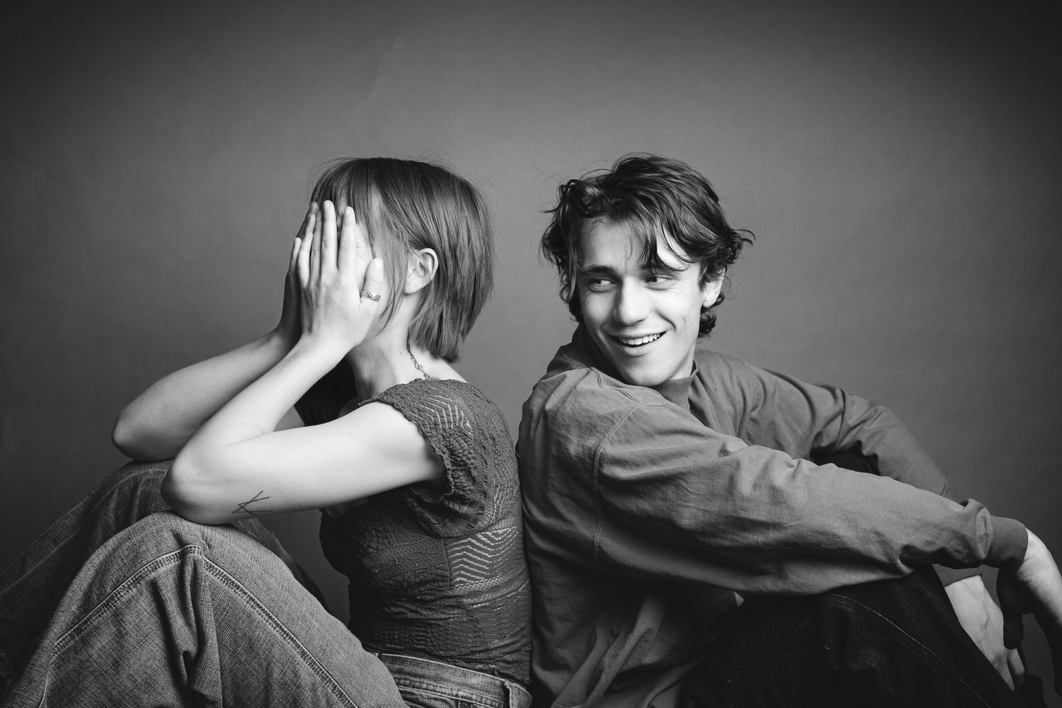 Black and white photo of two children sitting back to back, boy smiling, girl covering her face with hands.