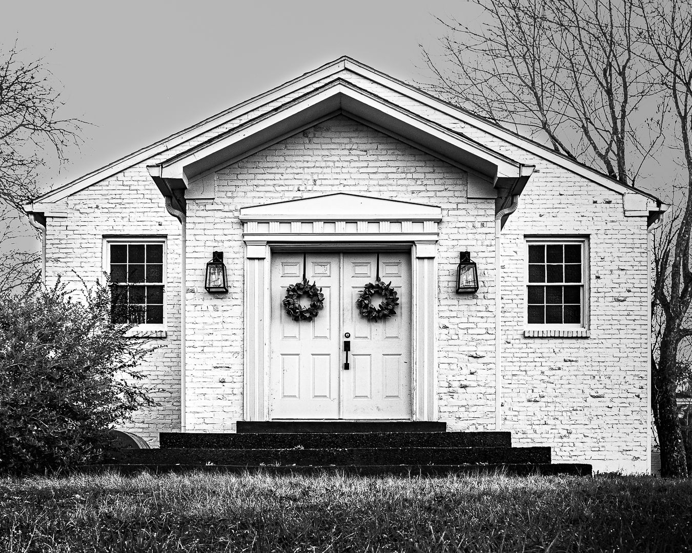 Black and white photo of a white brick church with double doors adorned with Christmas wreaths and lanterns.