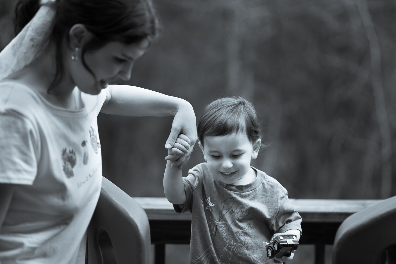 Mother and young son smiling together on outdoor deck, boy holding small toy in hand.