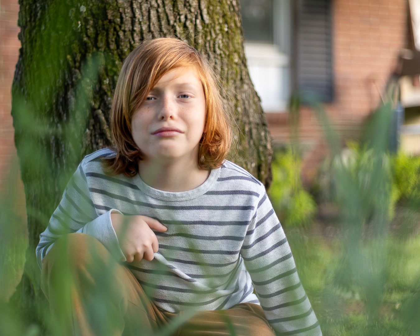 Young child with reddish hair sitting against a tree trunk in a garden wearing a striped shirt.