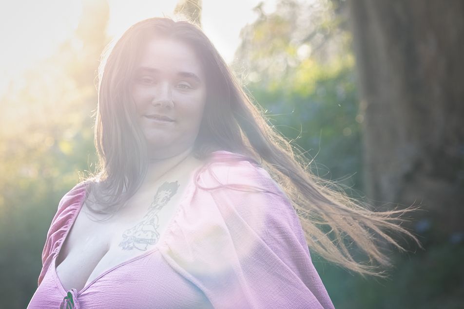 Woman with long hair in pink cape outdoors, bathed in warm sunlight with green trees in background.
