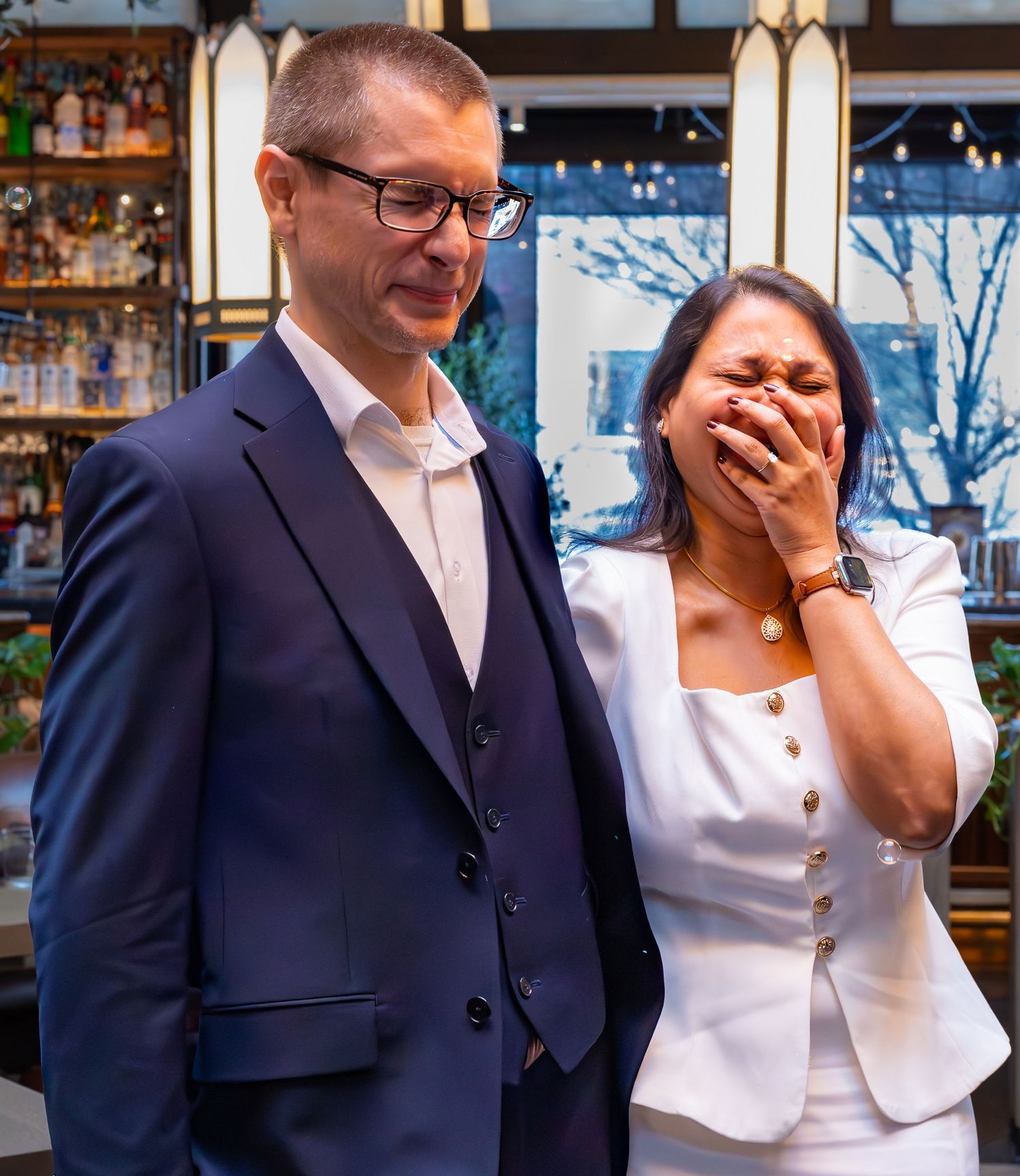 A man in a navy suit and a woman in a white blazer share an emotional moment at an elegant bar restaurant.