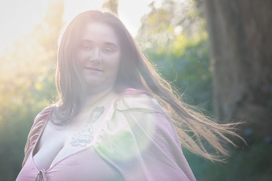 Young woman with long flowing hair smiling outdoors in soft sunlight with green trees in background.
