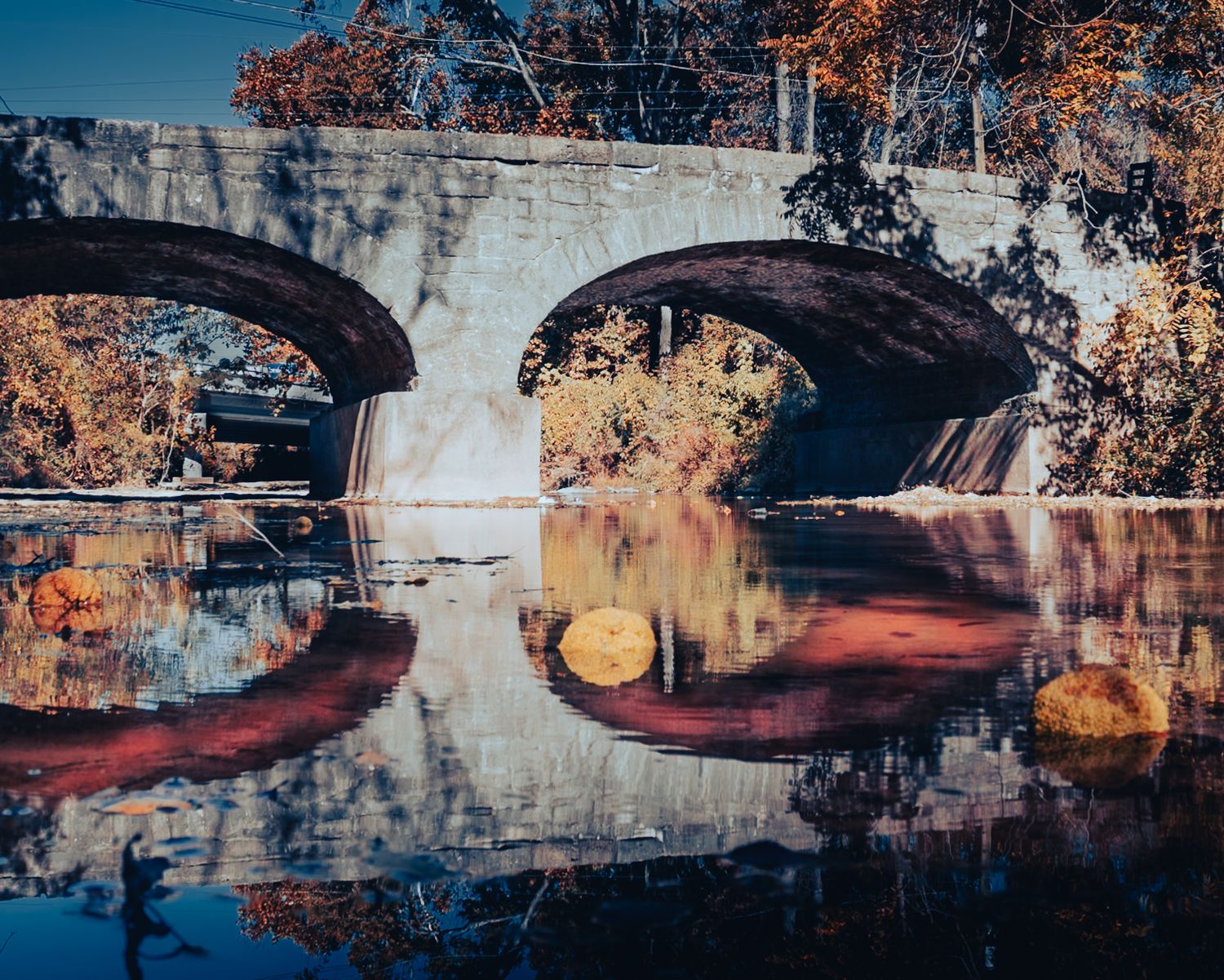 Stone bridge with twin arches reflecting on calm water surrounded by autumn trees in warm fall colors.