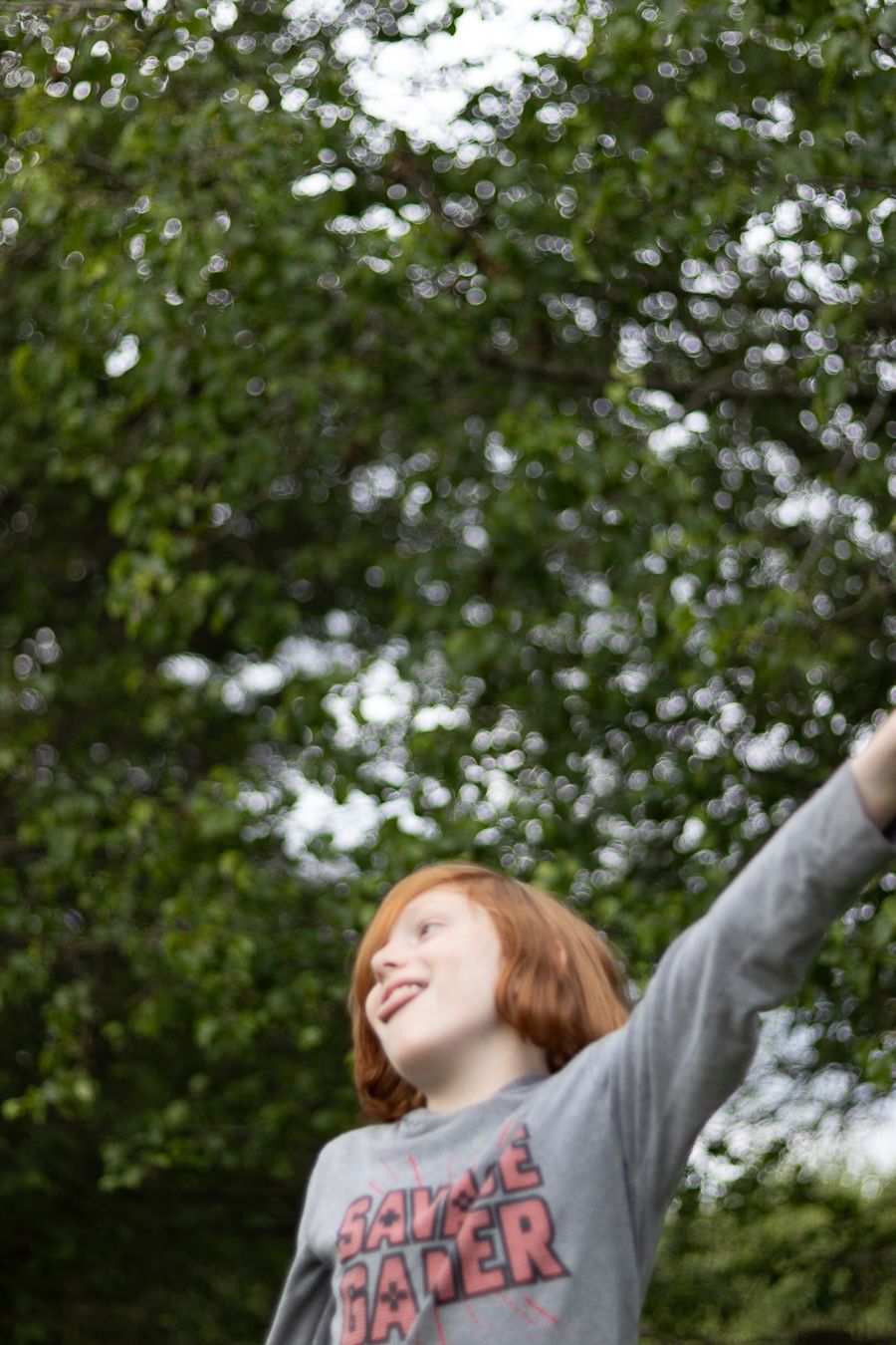 Happy redhead child outdoors reaching up toward green trees while wearing a graphic sweatshirt.
