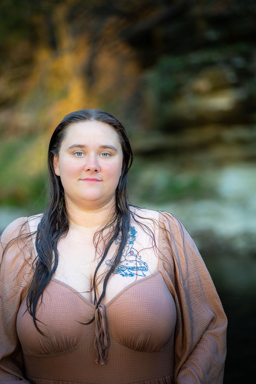 Woman with wet dark hair and blue eyes wearing a dress near a rocky waterfall outdoors.
