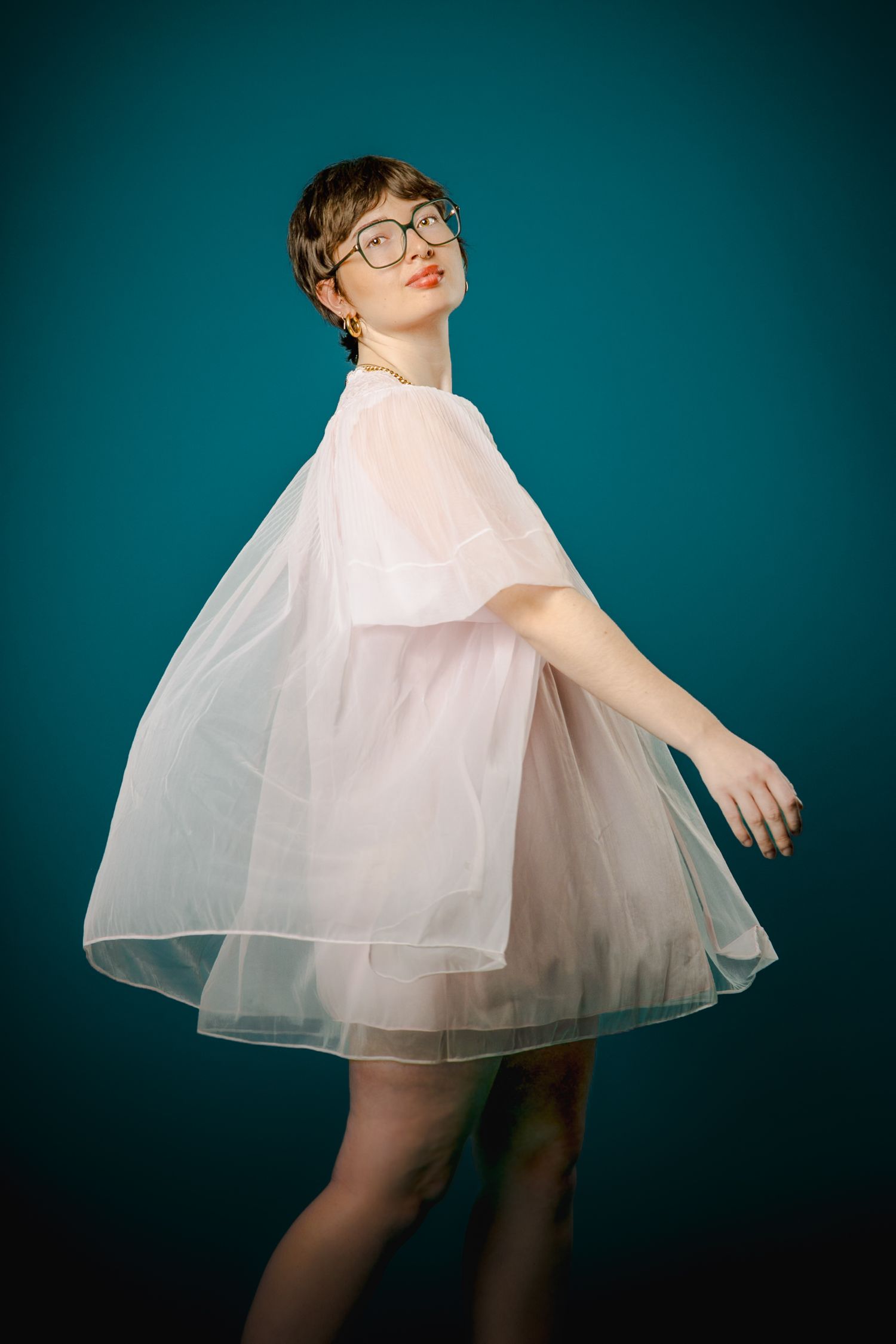 Young woman with short hair and glasses posing in a sheer white tulle dress against a teal background.