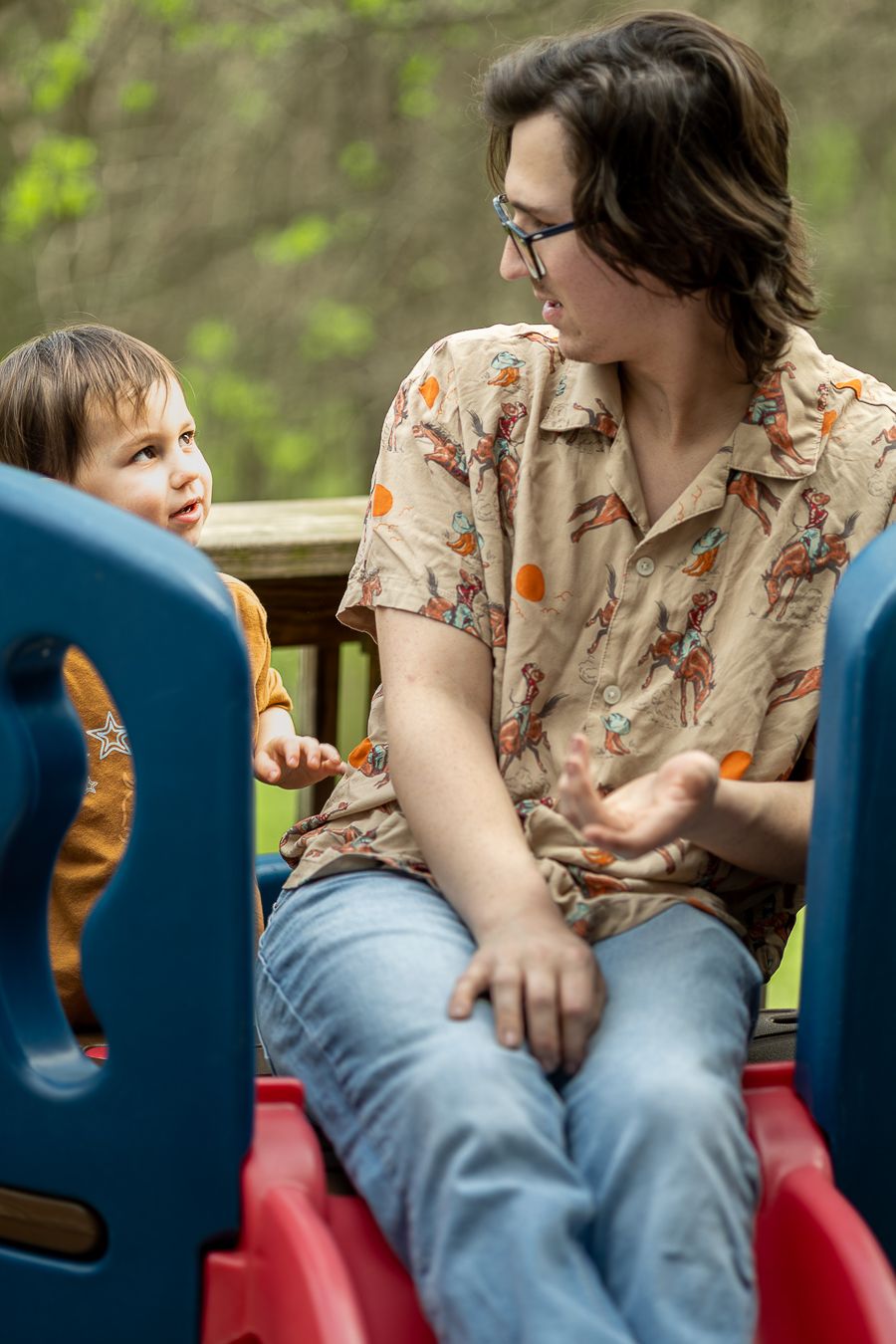 Father in patterned shirt talking with young toddler son on colorful outdoor playground.