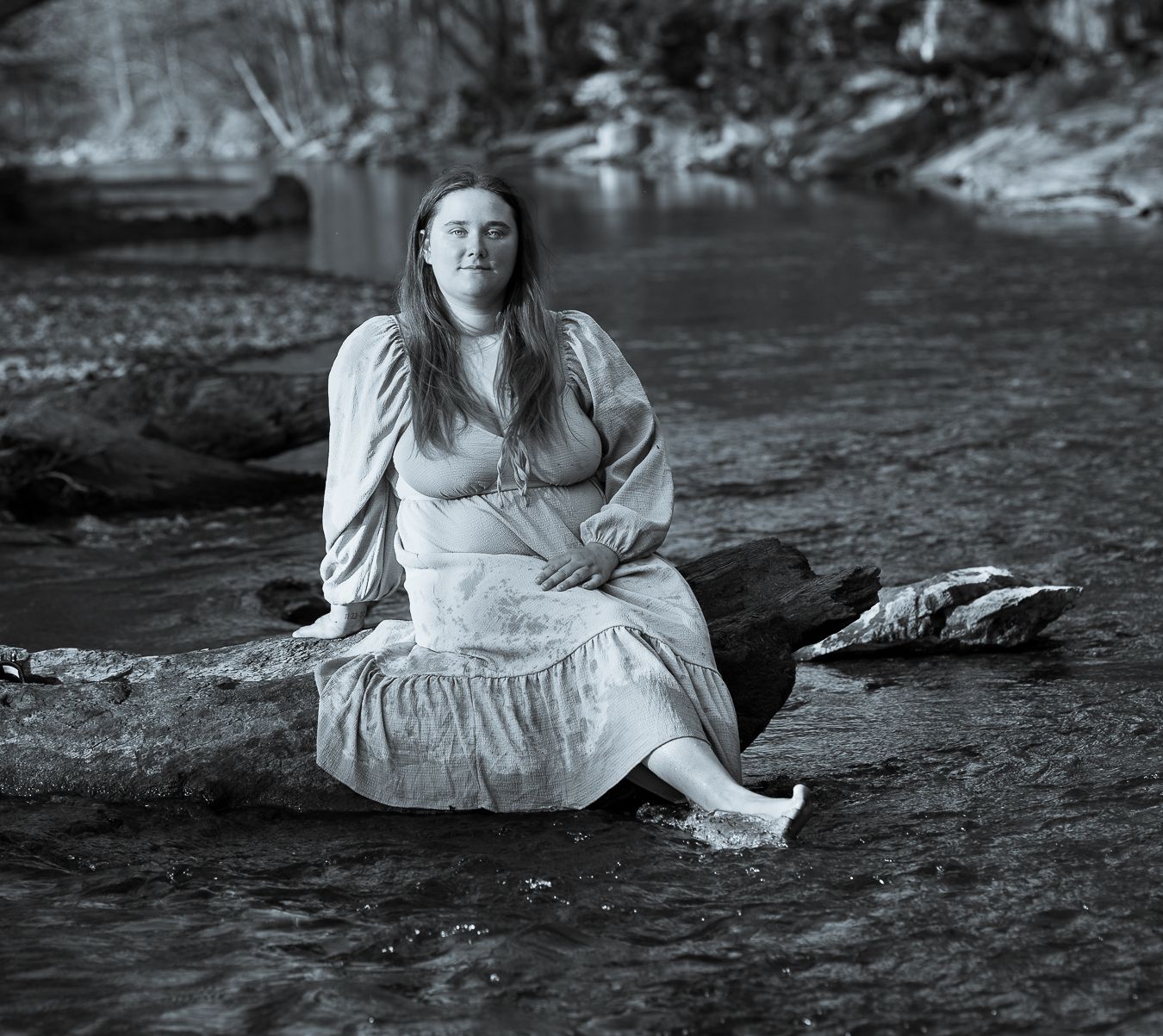 Black and white photo of a woman in a flowing dress sitting on a rock in a shallow stream.