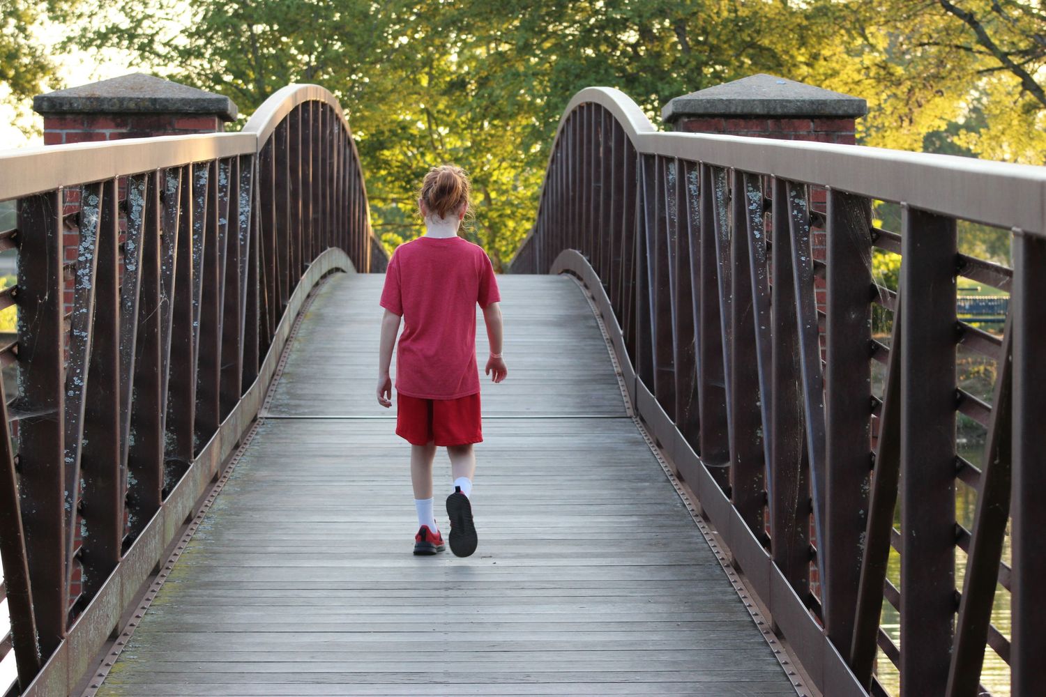 A boy in red walks alone across a wooden footbridge surrounded by autumn trees.