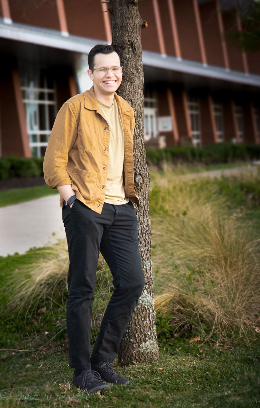 Smiling young man in mustard yellow jacket leaning against tree trunk outside modern building on sunny day.
