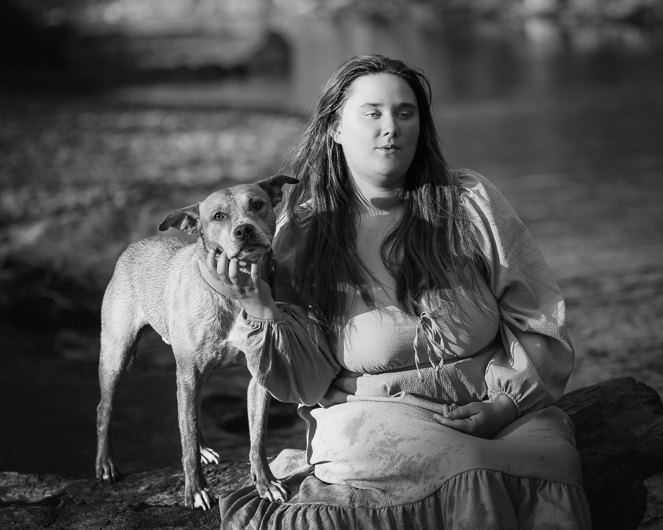 Black and white portrait of a woman sitting on rocks by water with a dog beside her.