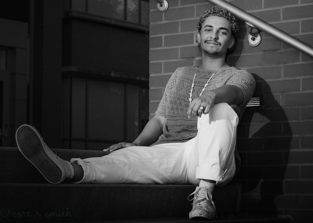 Young man in white pants and striped shirt sitting relaxed on outdoor steps against brick wall, black and white photo.