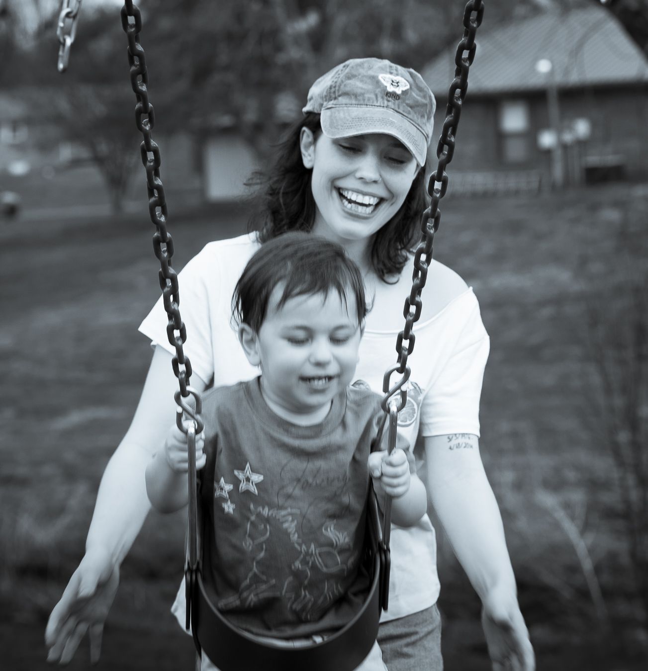 Black and white photo of smiling mother pushing young child on a playground swing.