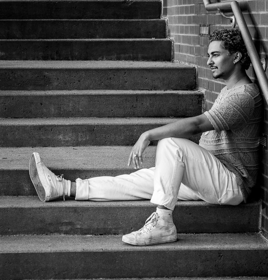 Young man in white pants and sneakers sitting on outdoor brick steps, posed thoughtfully in black and white photo.
