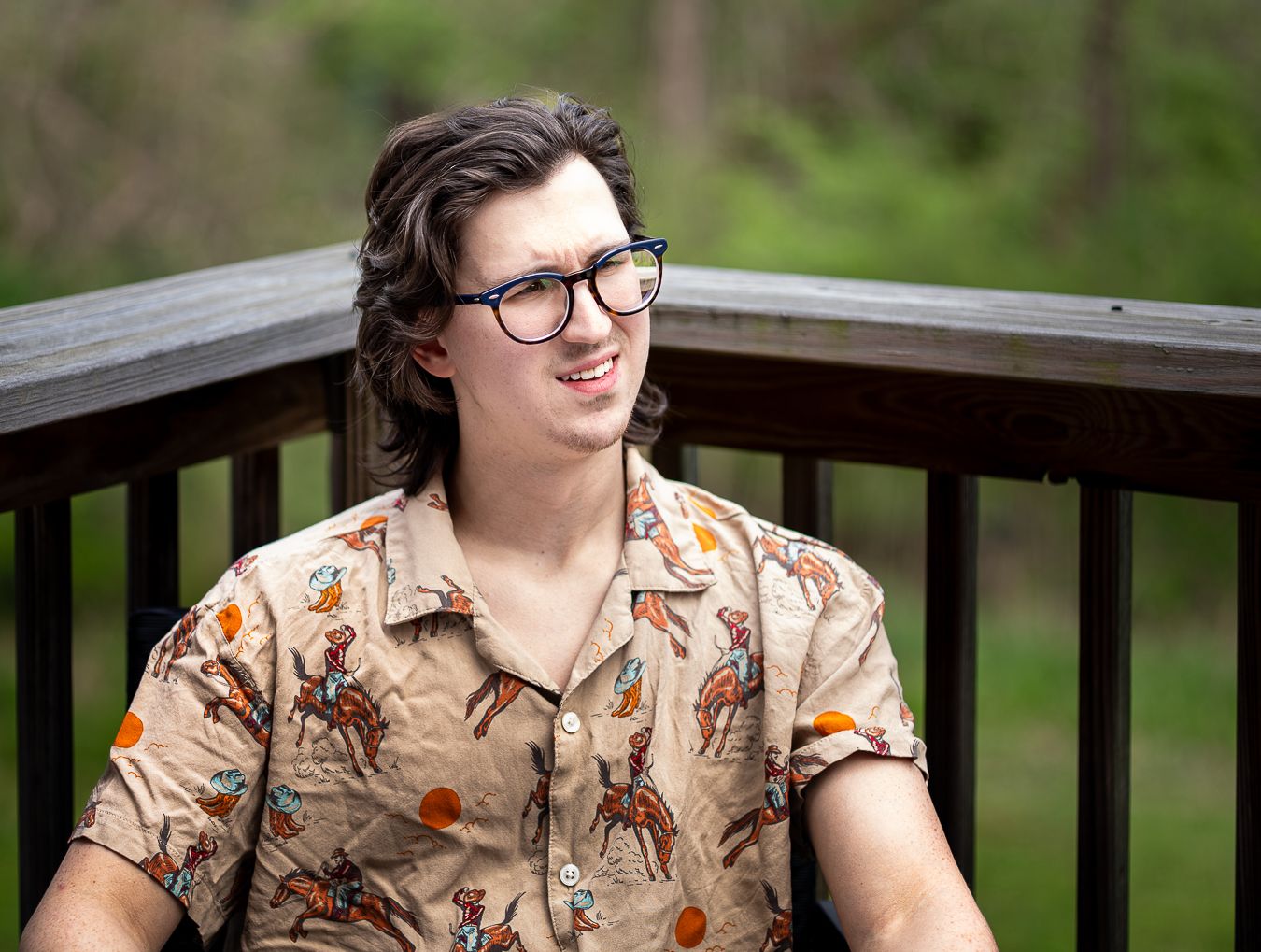 Young man with wavy hair and glasses smiling on a wooden deck wearing a cowboy print shirt.
