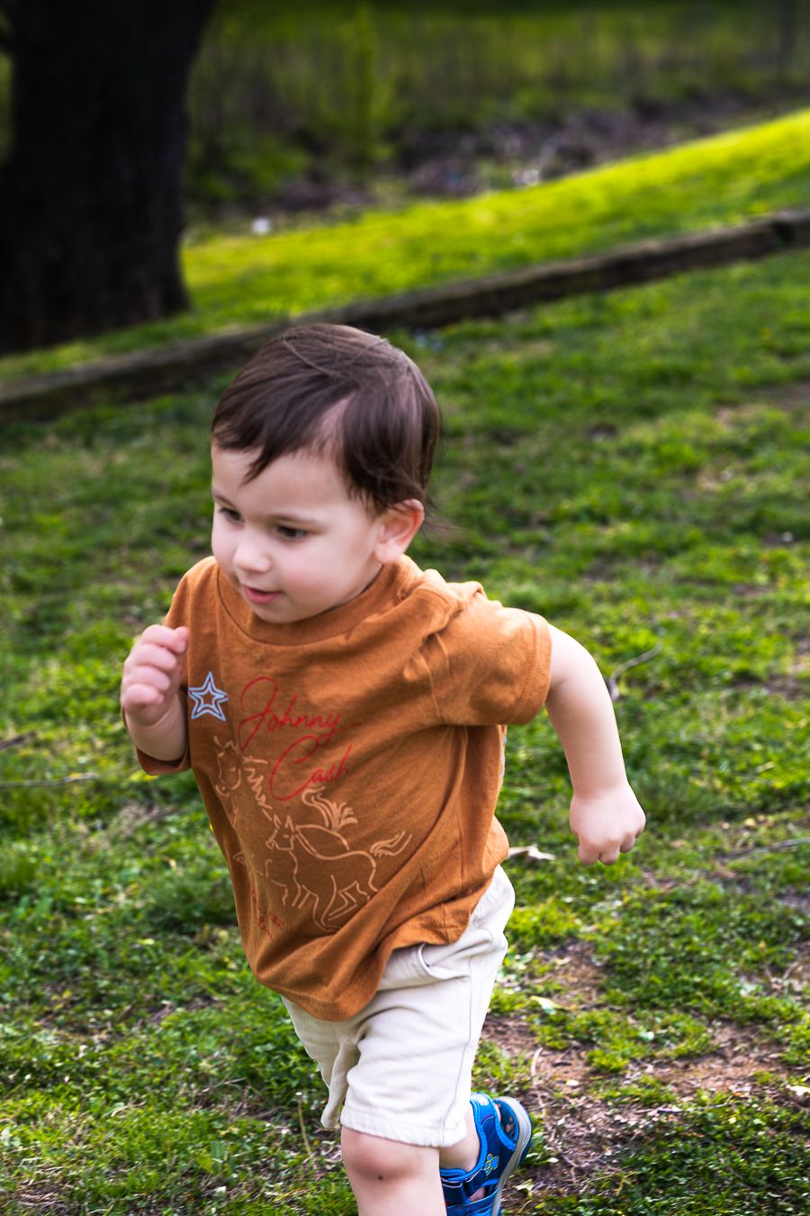 Energetic toddler boy in orange shirt running across green grass in a sunny park.