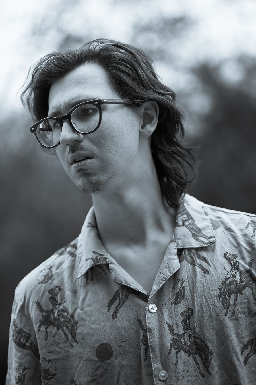 Black and white portrait of a young man with wavy hair and round glasses wearing a patterned shirt.