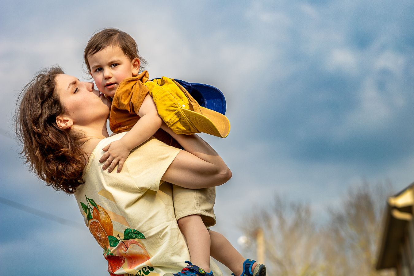 Mother lifting and kissing smiling toddler against a dramatic cloudy blue sky backdrop.