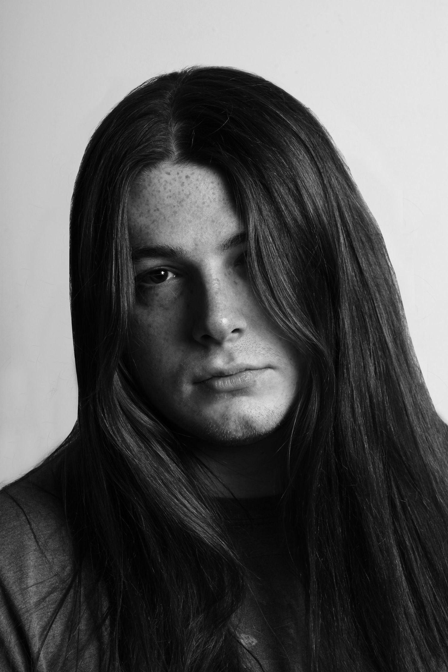 Black and white portrait of a young man with long straight hair and freckles looking at camera.