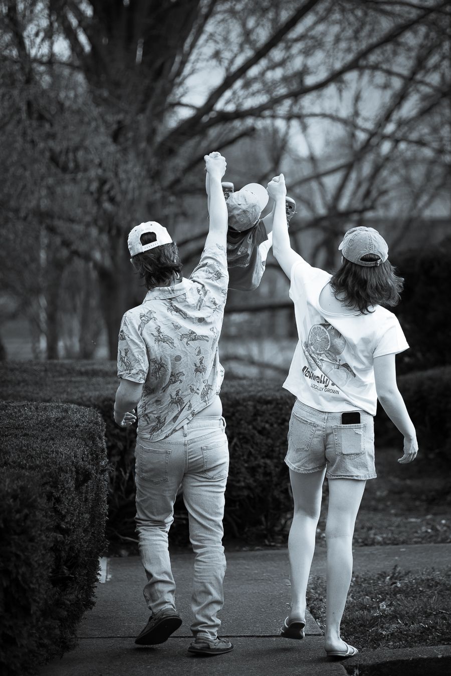 Black and white photo of parents lifting child by hands on a tree-lined sidewalk.