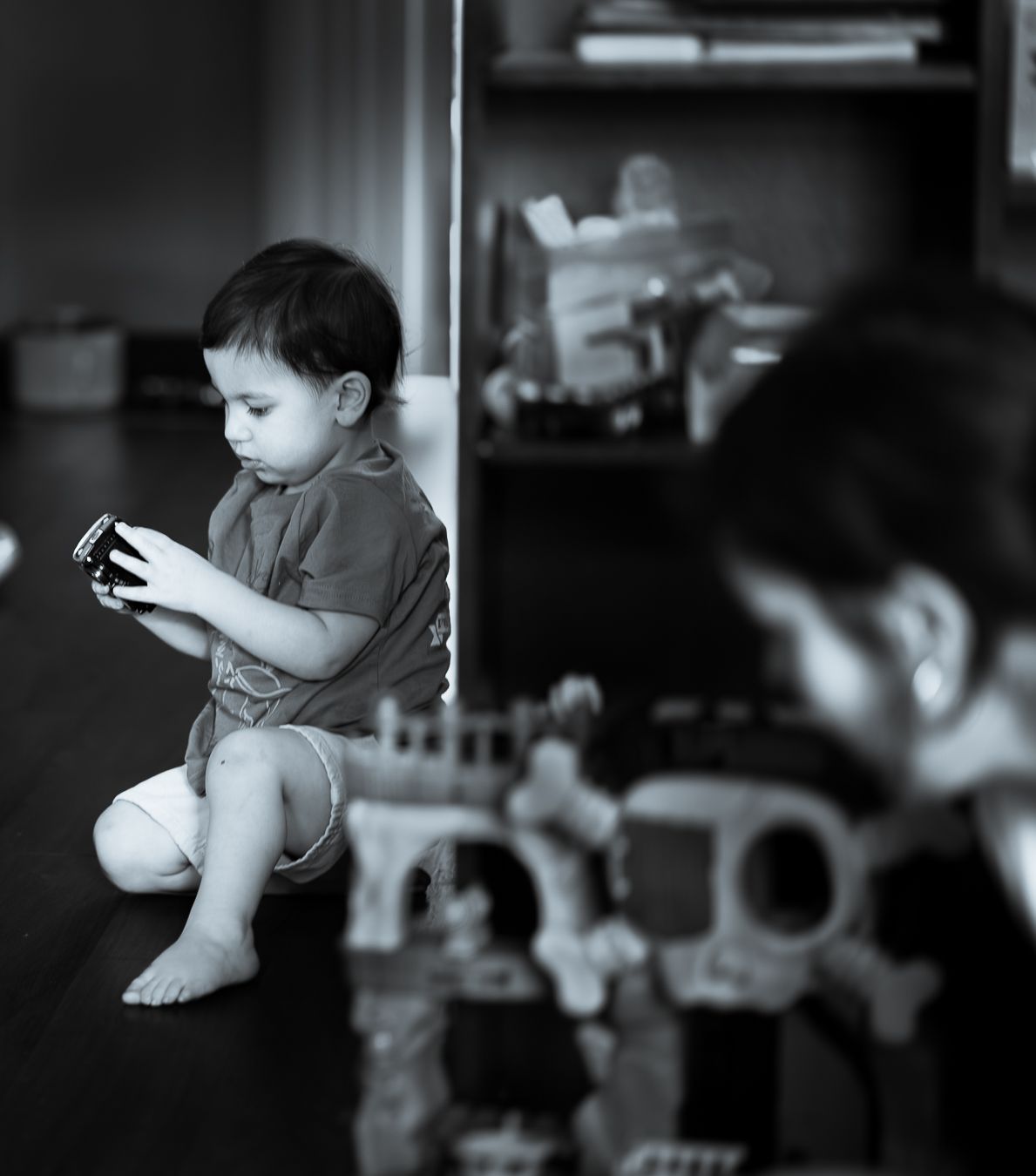 Young toddler sitting on floor playing with toy in a dimly lit room with toys in background.