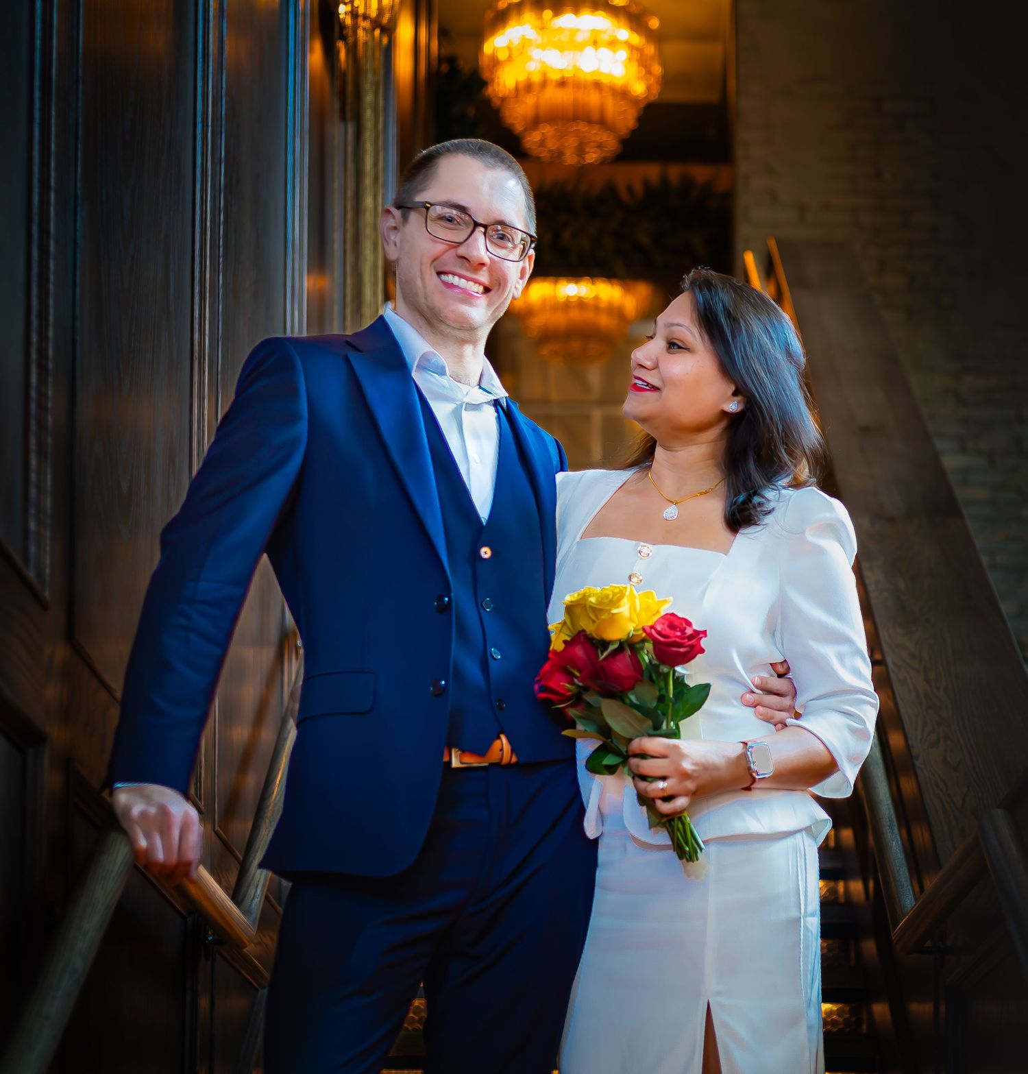 Smiling couple in wedding attire poses on staircase, groom in blue suit, bride holding red and yellow roses.