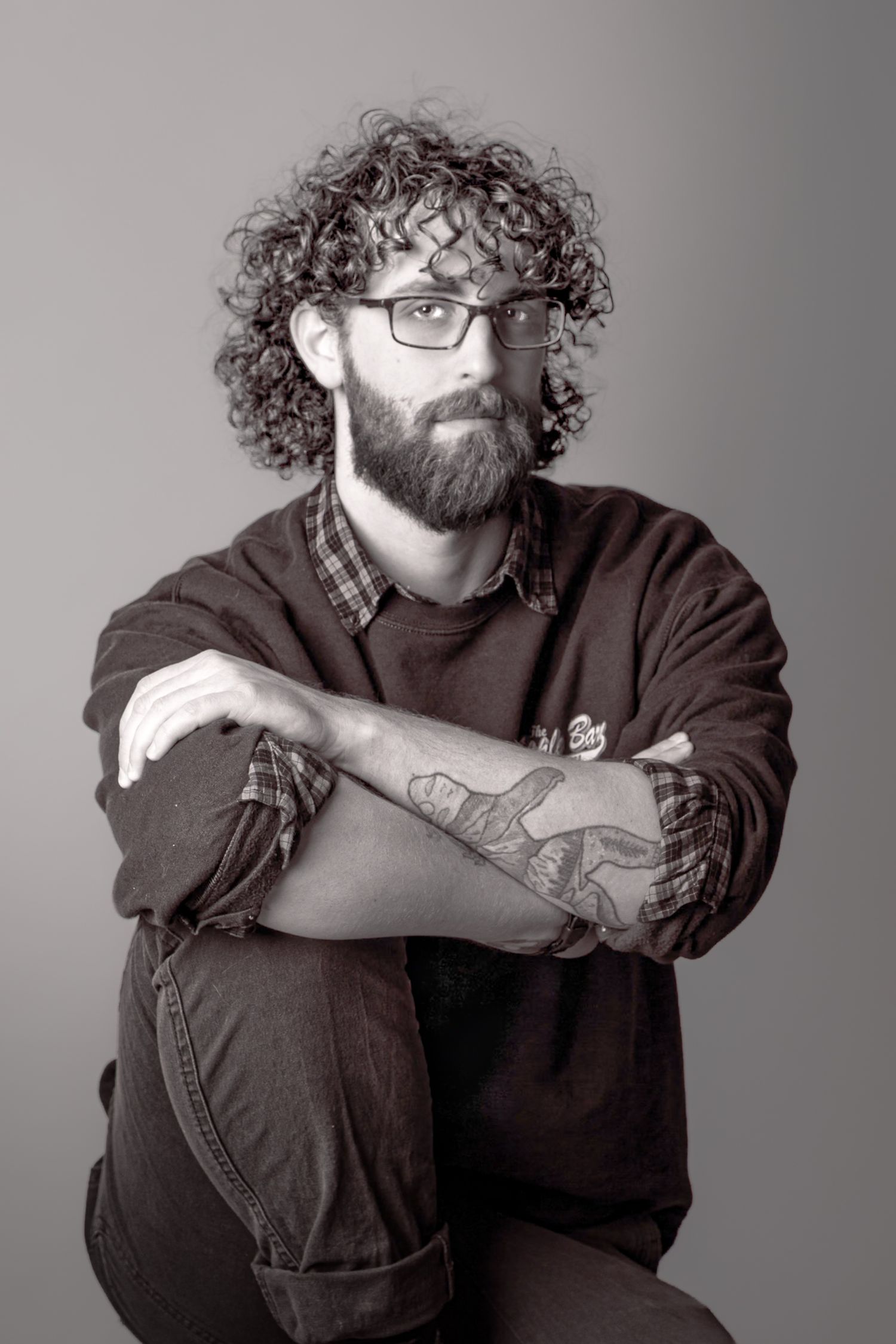 Black and white portrait of bearded curly-haired man with glasses and tattoos sitting with arms crossed.