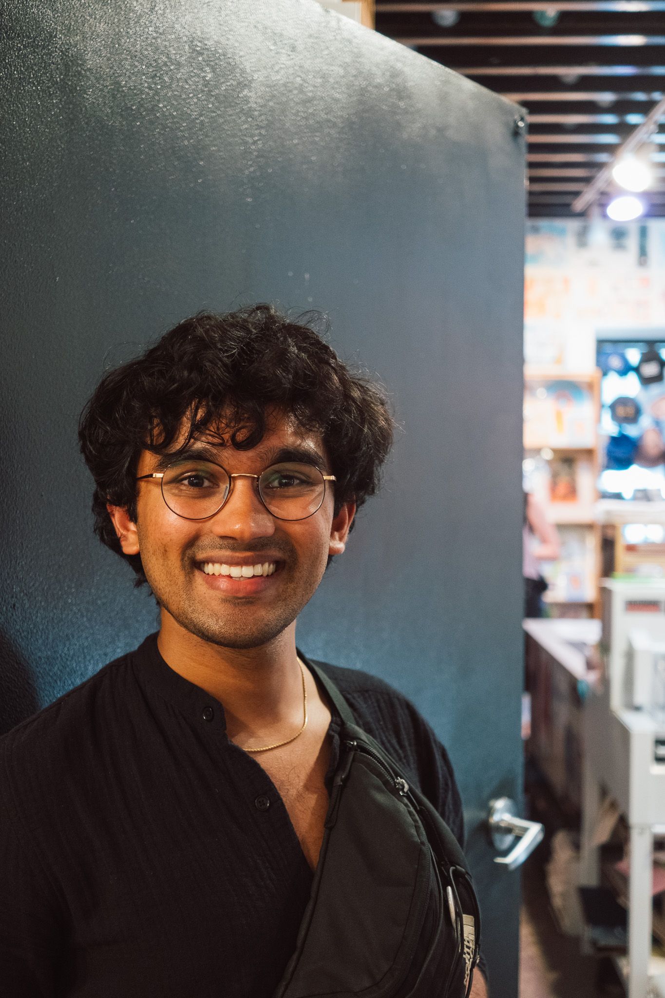 Smiling young man with curly hair and round glasses leaning against a dark wall in a bookstore.