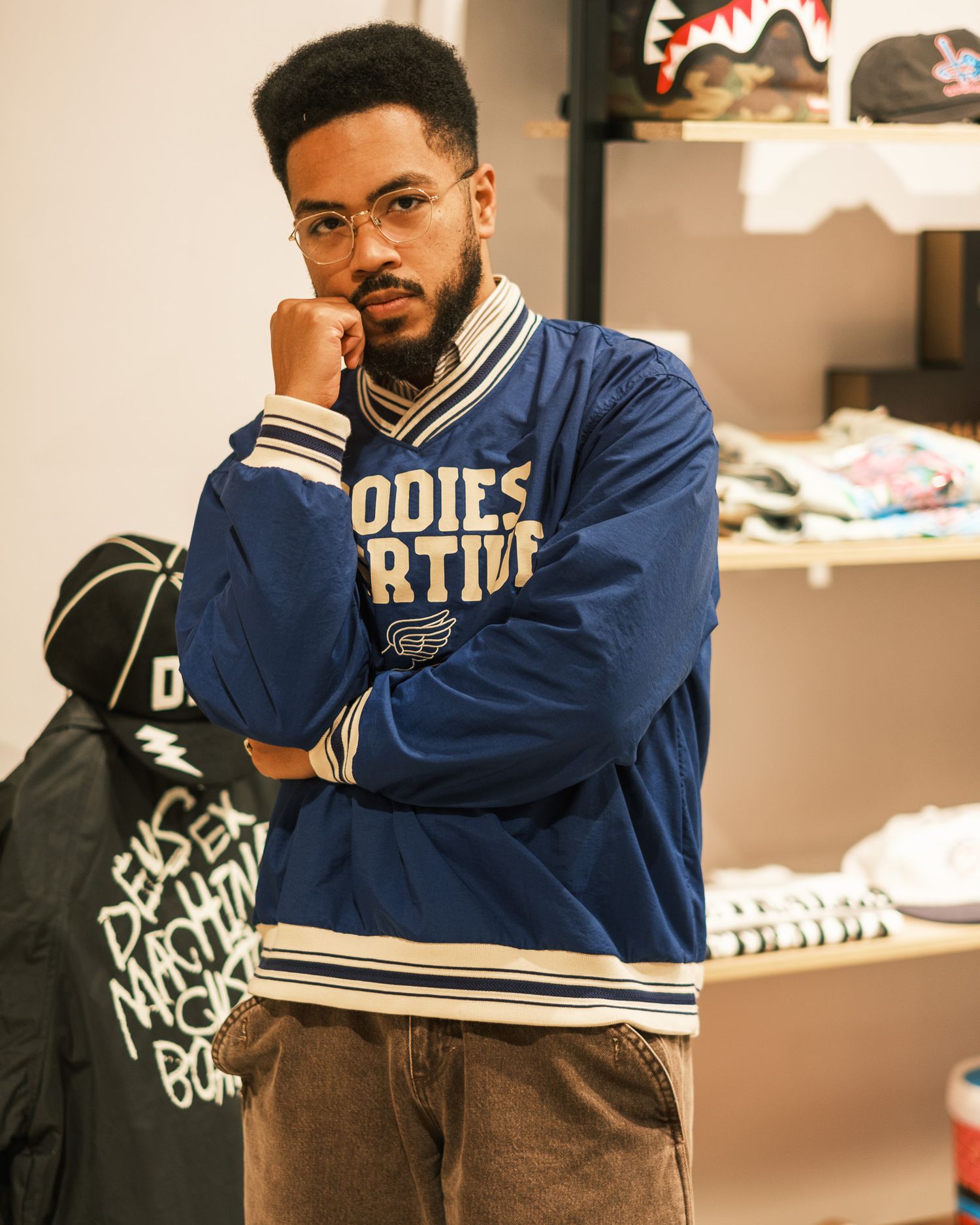 Young man in a blue varsity jacket posing thoughtfully inside a streetwear clothing store.