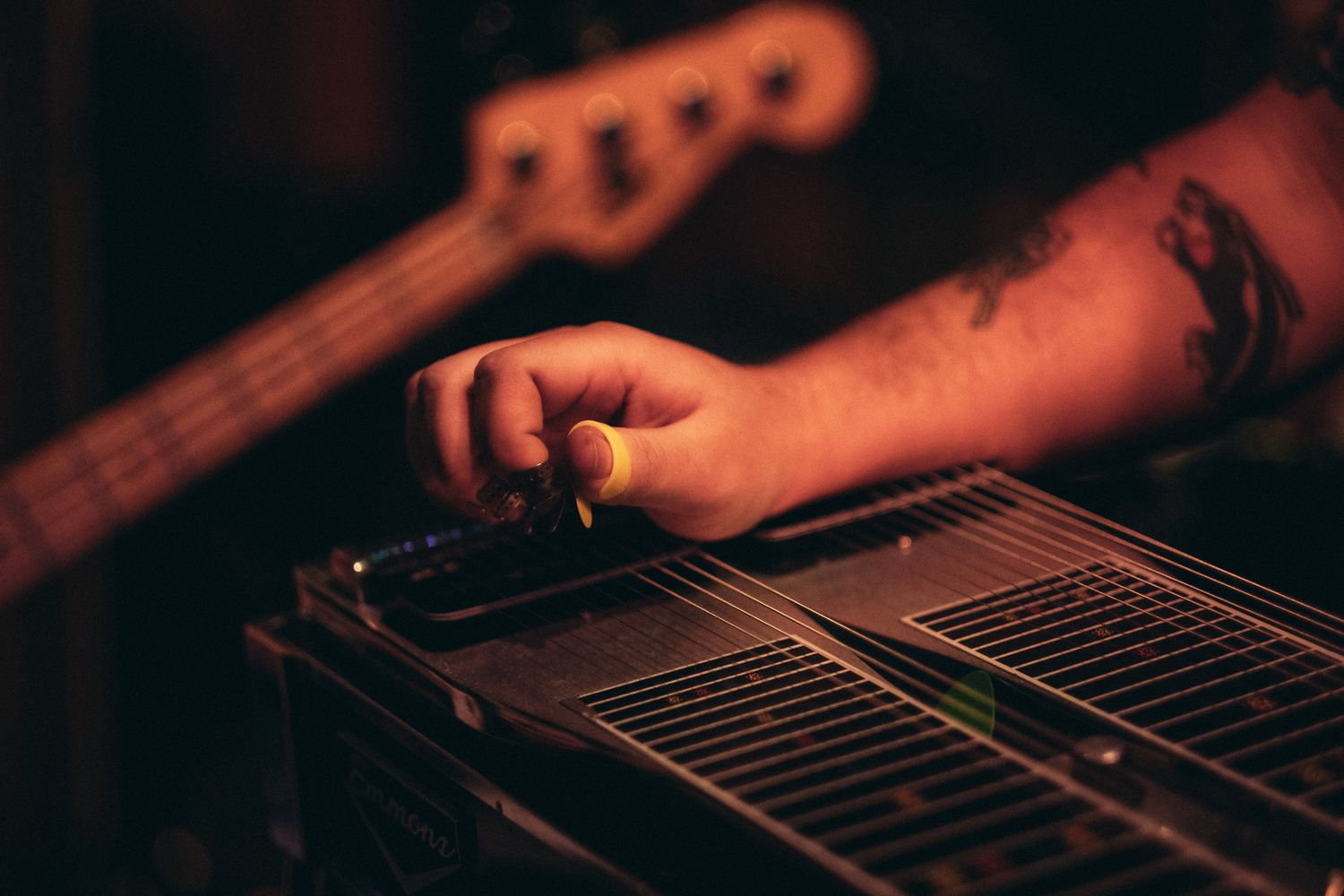 A tattooed hand plays a lap steel guitar with a bass guitar visible in the background.