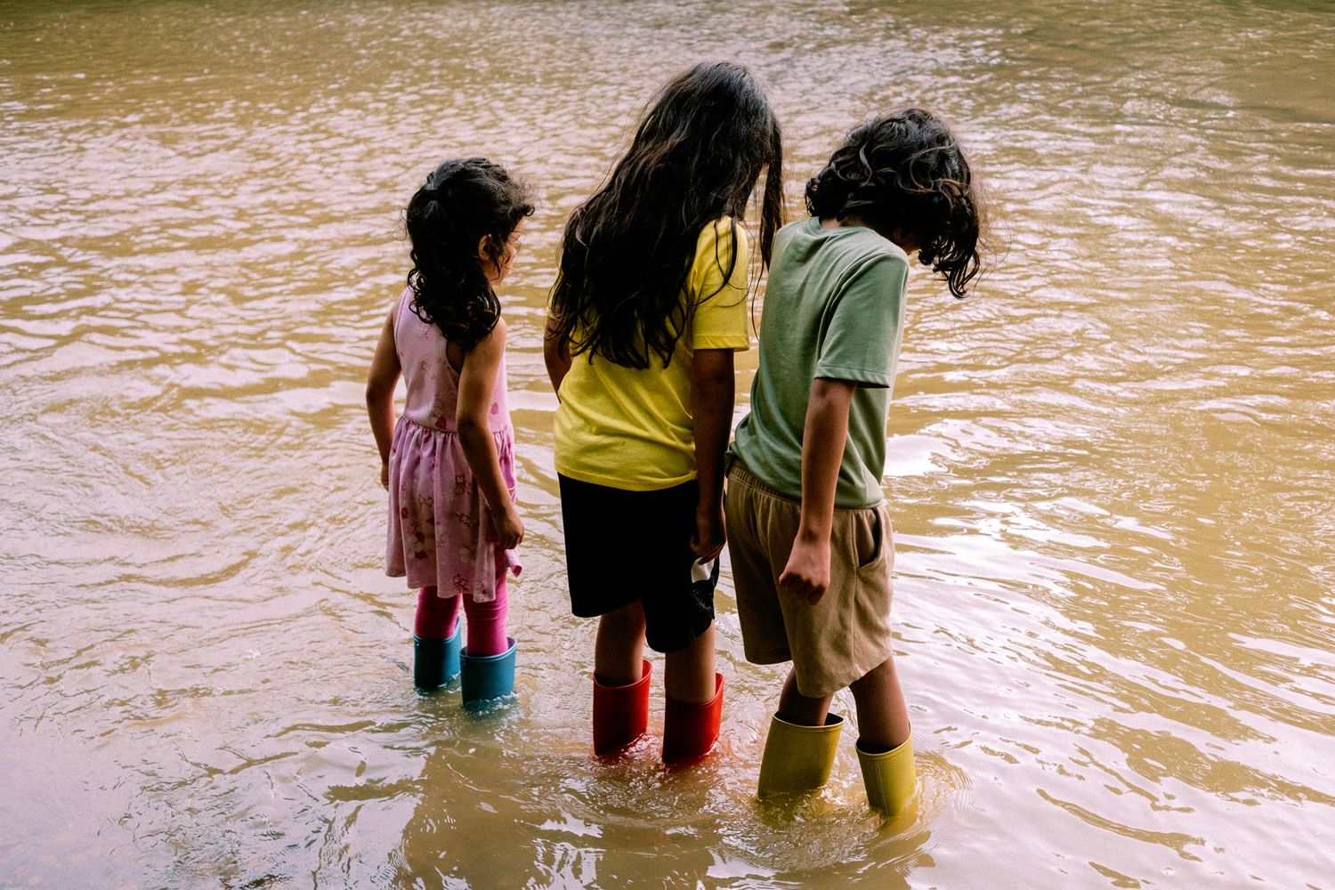 Three children standing in shallow muddy water wearing colorful rain boots, viewed from behind.