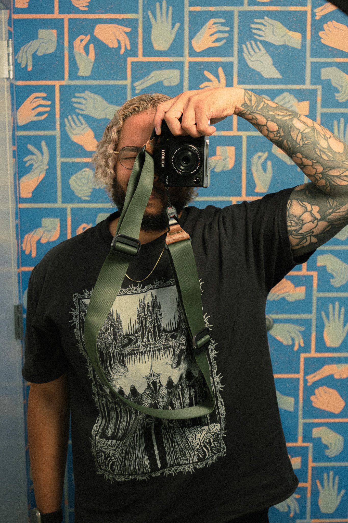 Tattooed man with curly hair taking mirror selfie with camera in front of blue hand-print wall.