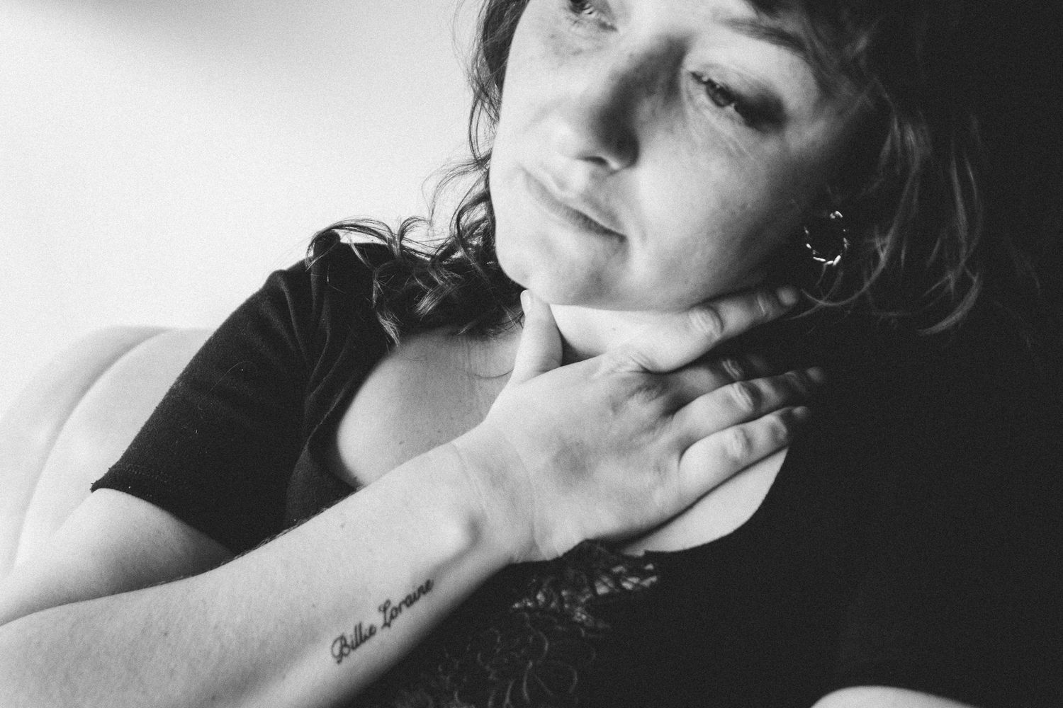 Black and white close-up of a woman touching her neck with a script tattoo on her forearm.