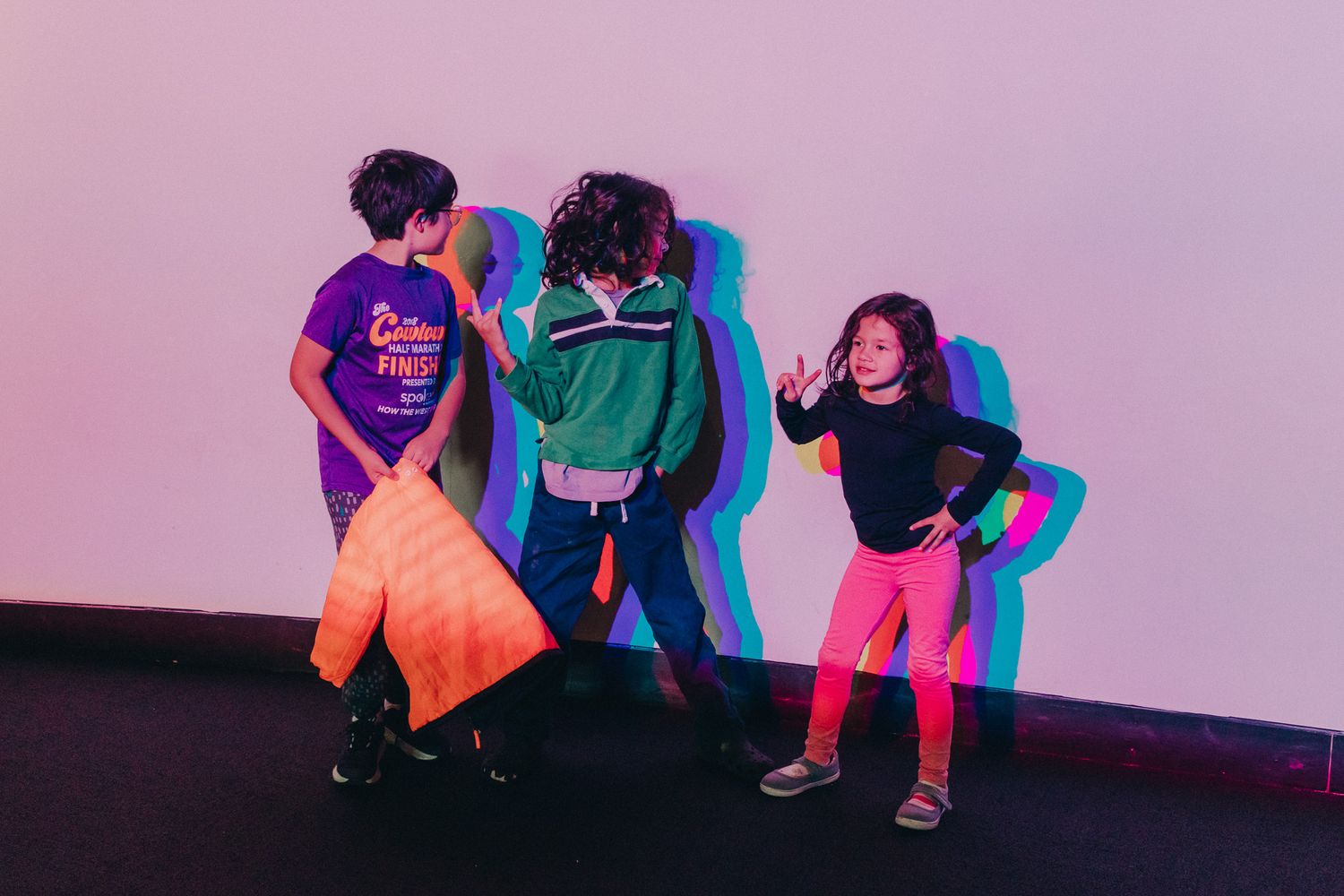 Three children playing and dancing under colorful neon lights against a pink wall indoors.