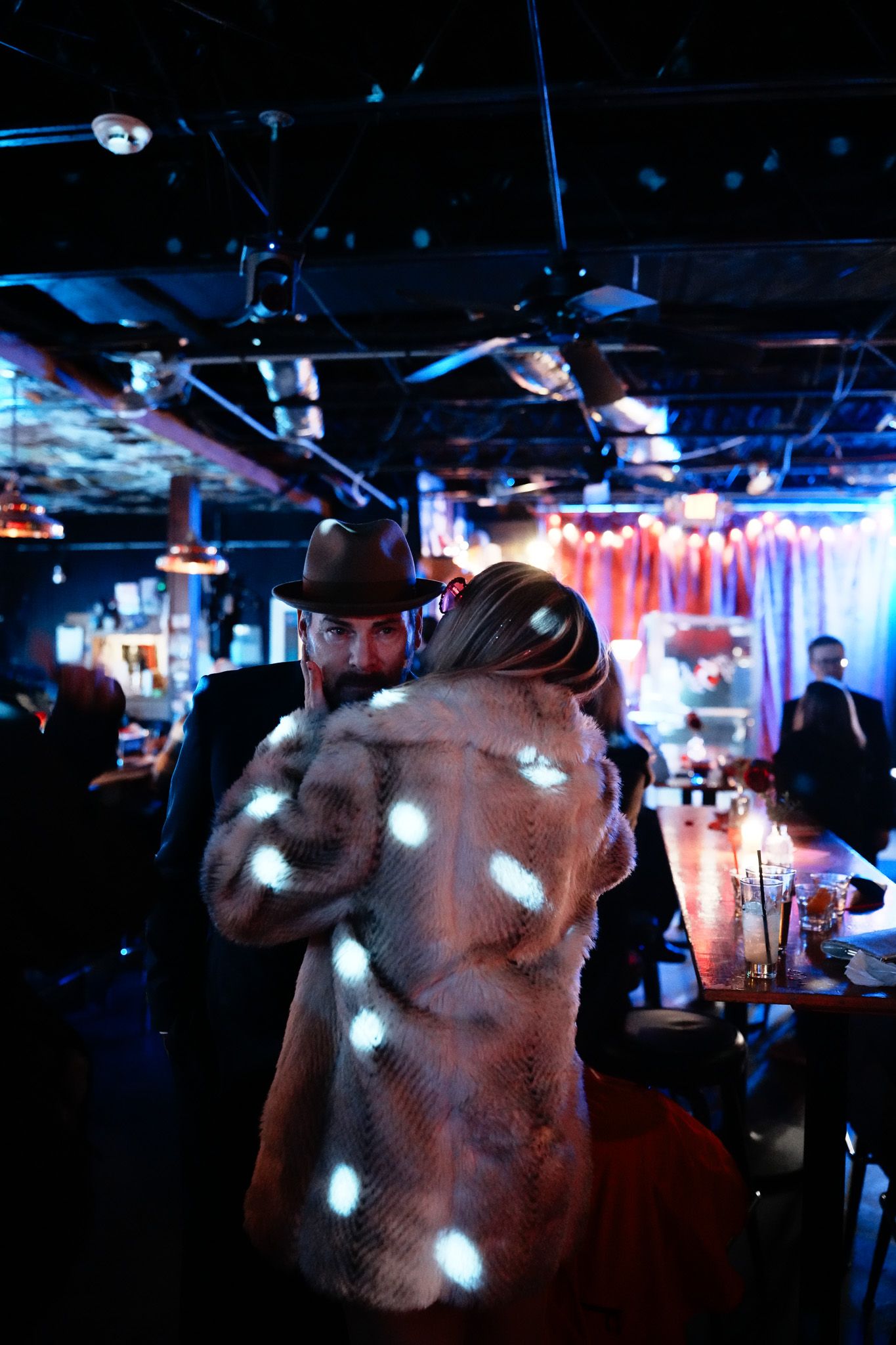 Person in fur coat and cowboy hat dancing in a vibrant blue-lit bar or nightclub venue.