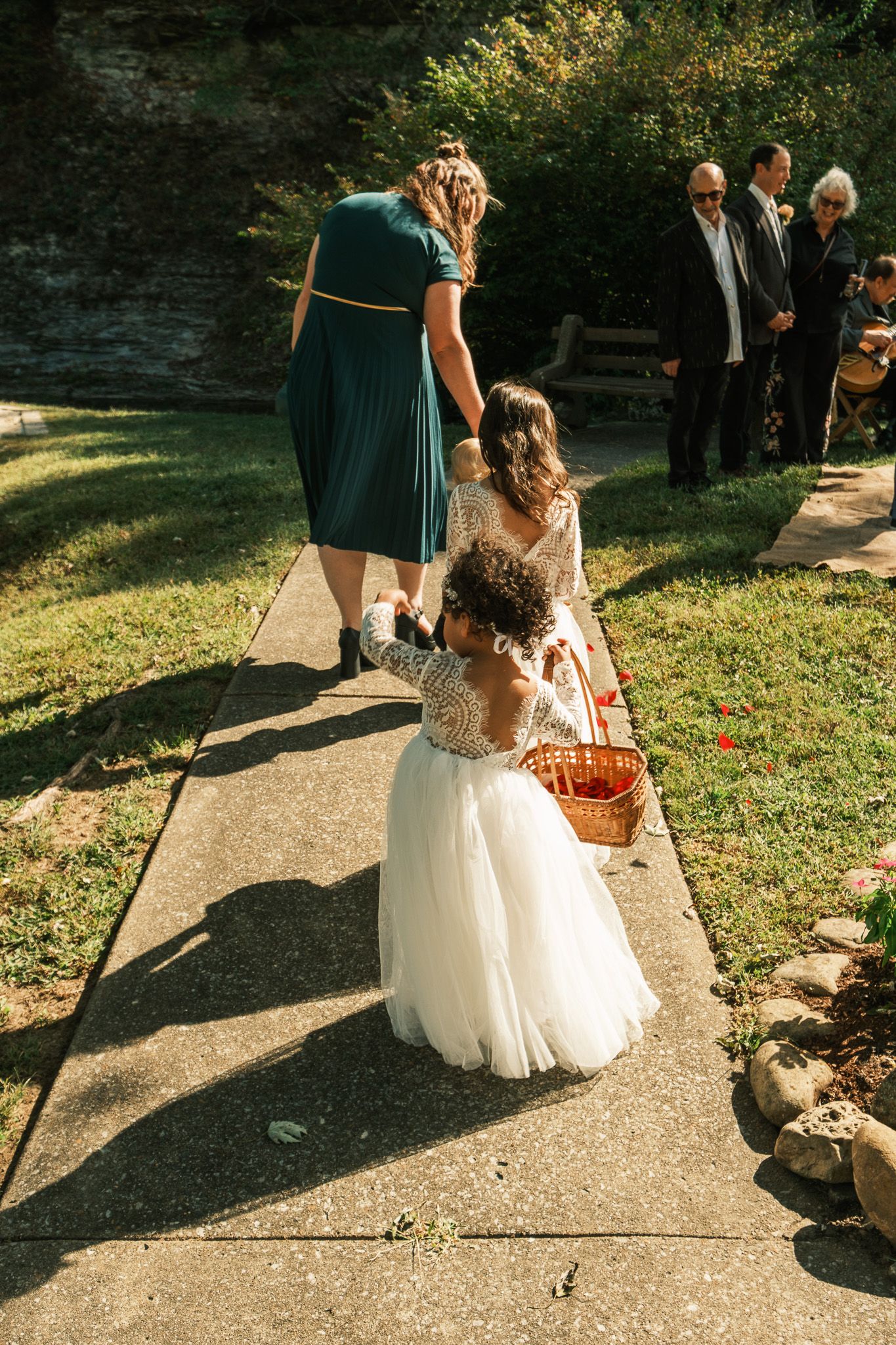 Flower girl in white tulle dress tosses petals on the aisle assisted by bridesmaid in teal dress.