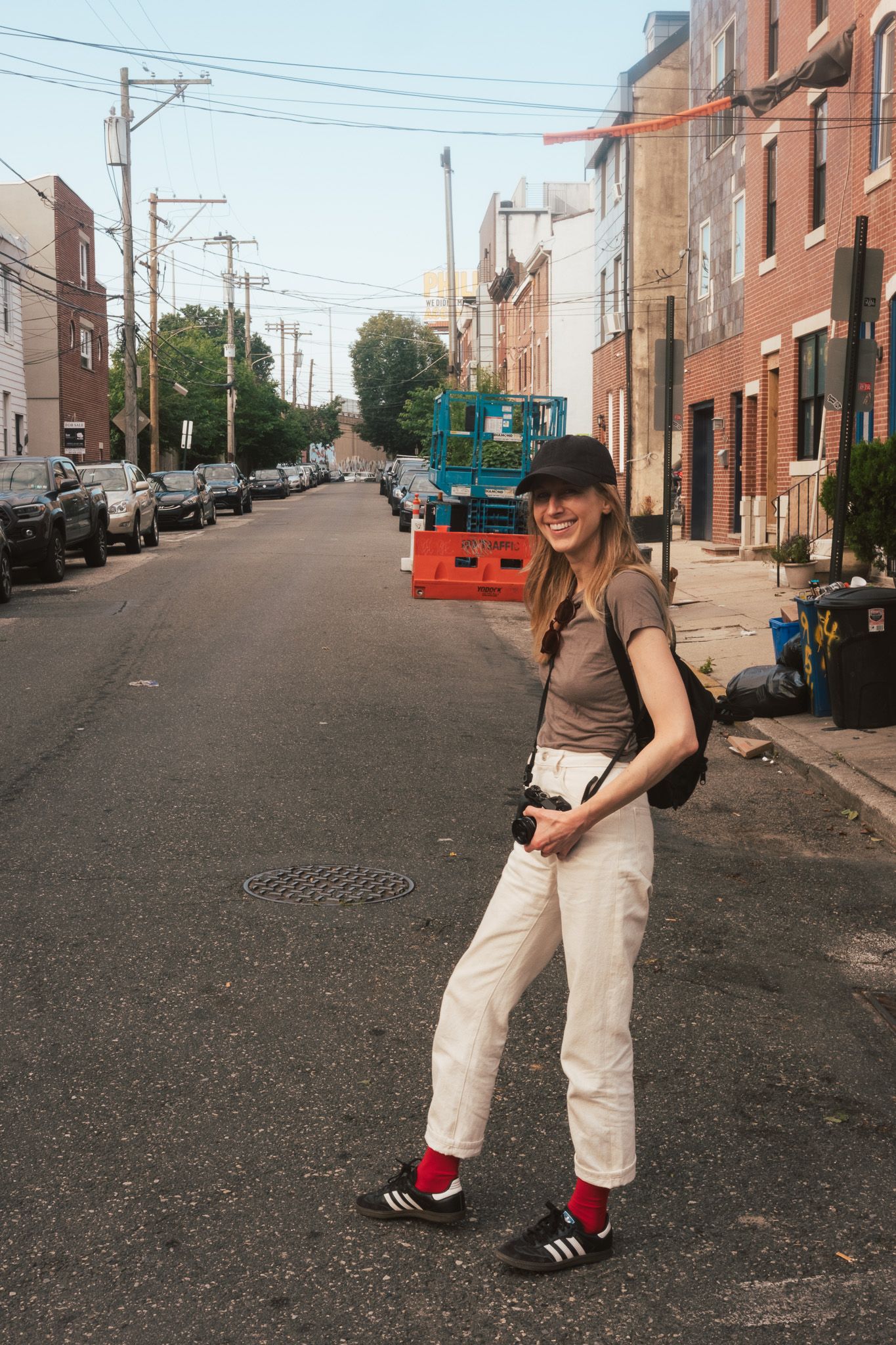 Young woman in white jeans and brown t-shirt posing on an urban street holding a camera, wearing a black cap.