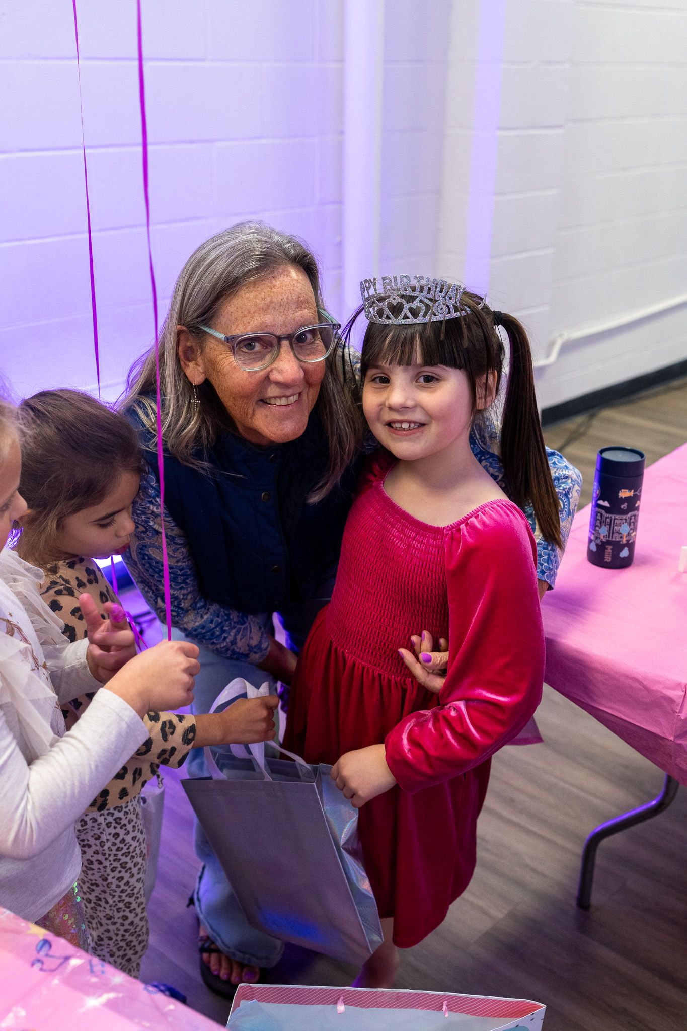 Grandmother and birthday girl in red dress with tiara smile together at a pink-themed party celebration.