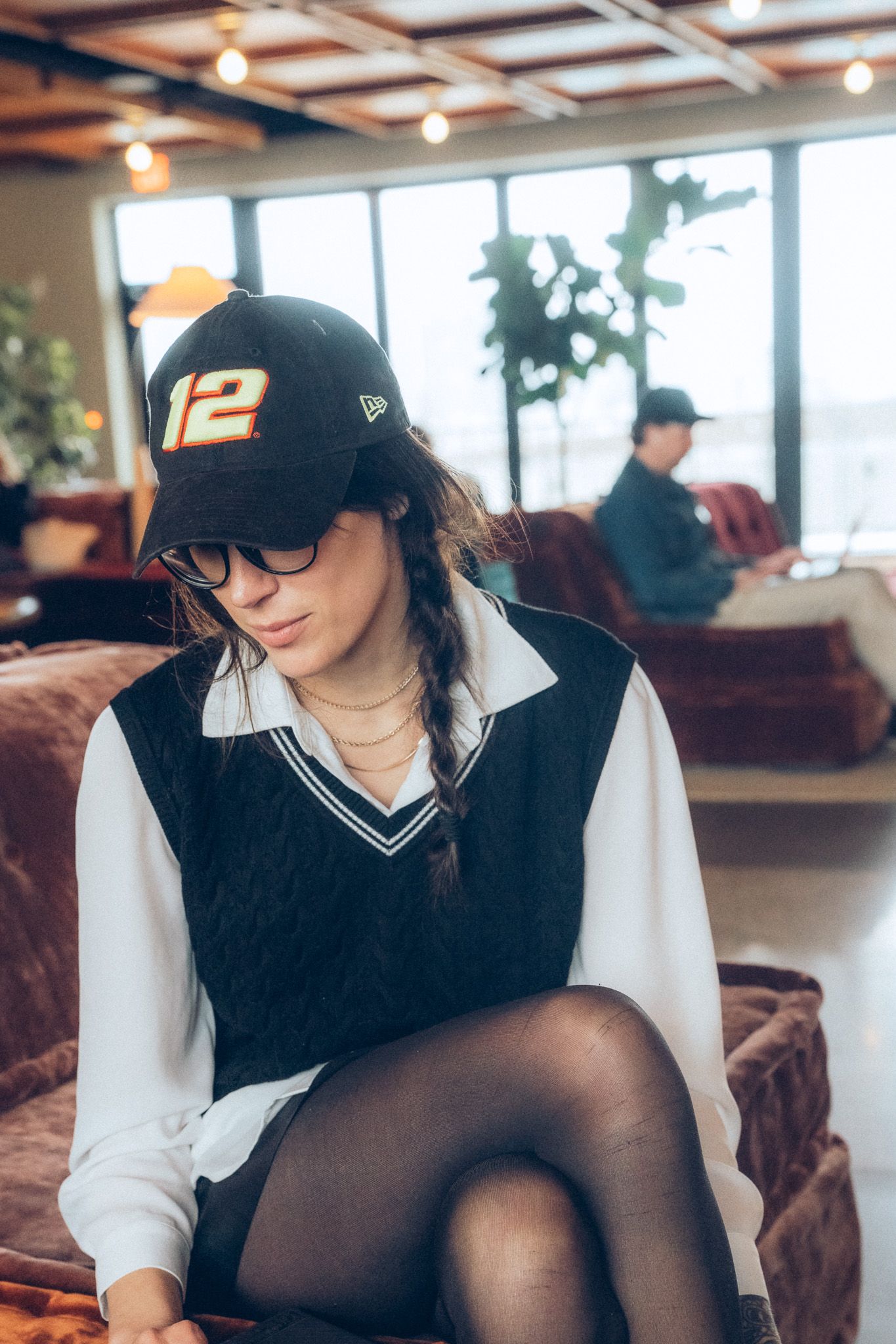 Young woman in a number 12 cap and glasses sitting on a velvet chair in a cozy cafe.