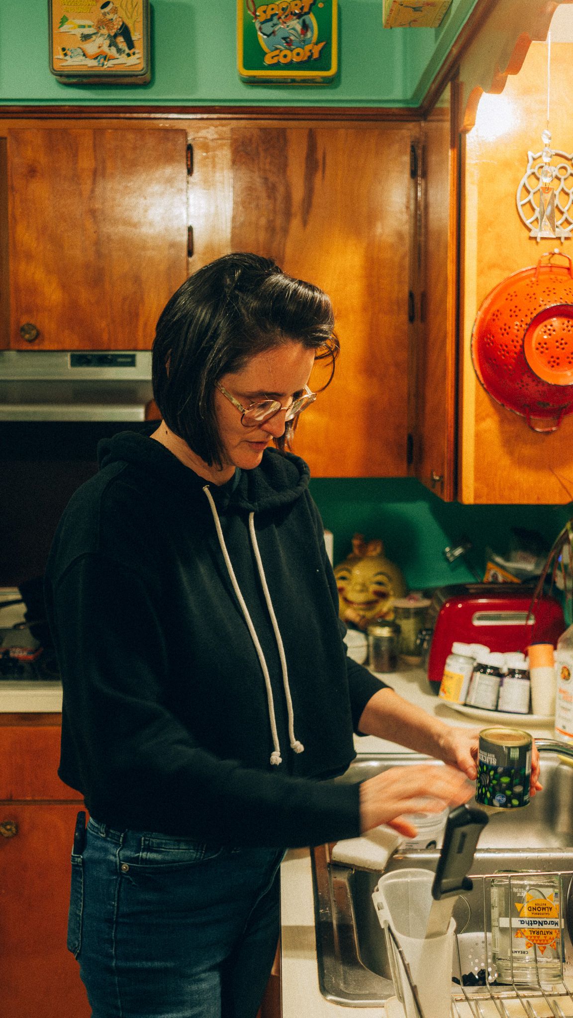 Woman in black hoodie and glasses cooking in a retro kitchen with wooden cabinets and green walls.