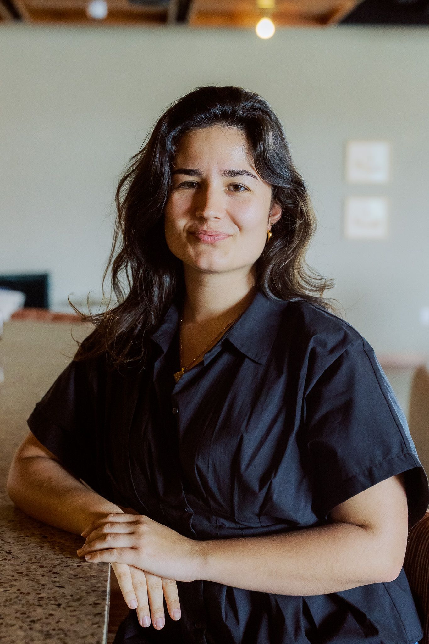 Young woman with dark wavy hair in a black dress smiling while leaning on a counter indoors.
