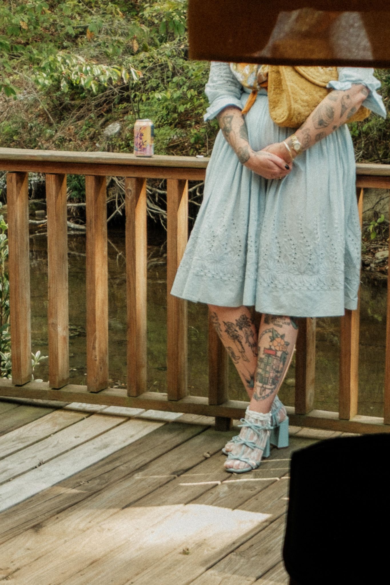 Woman in light blue eyelet dress with extensive leg tattoos wearing blue heels on wooden deck.