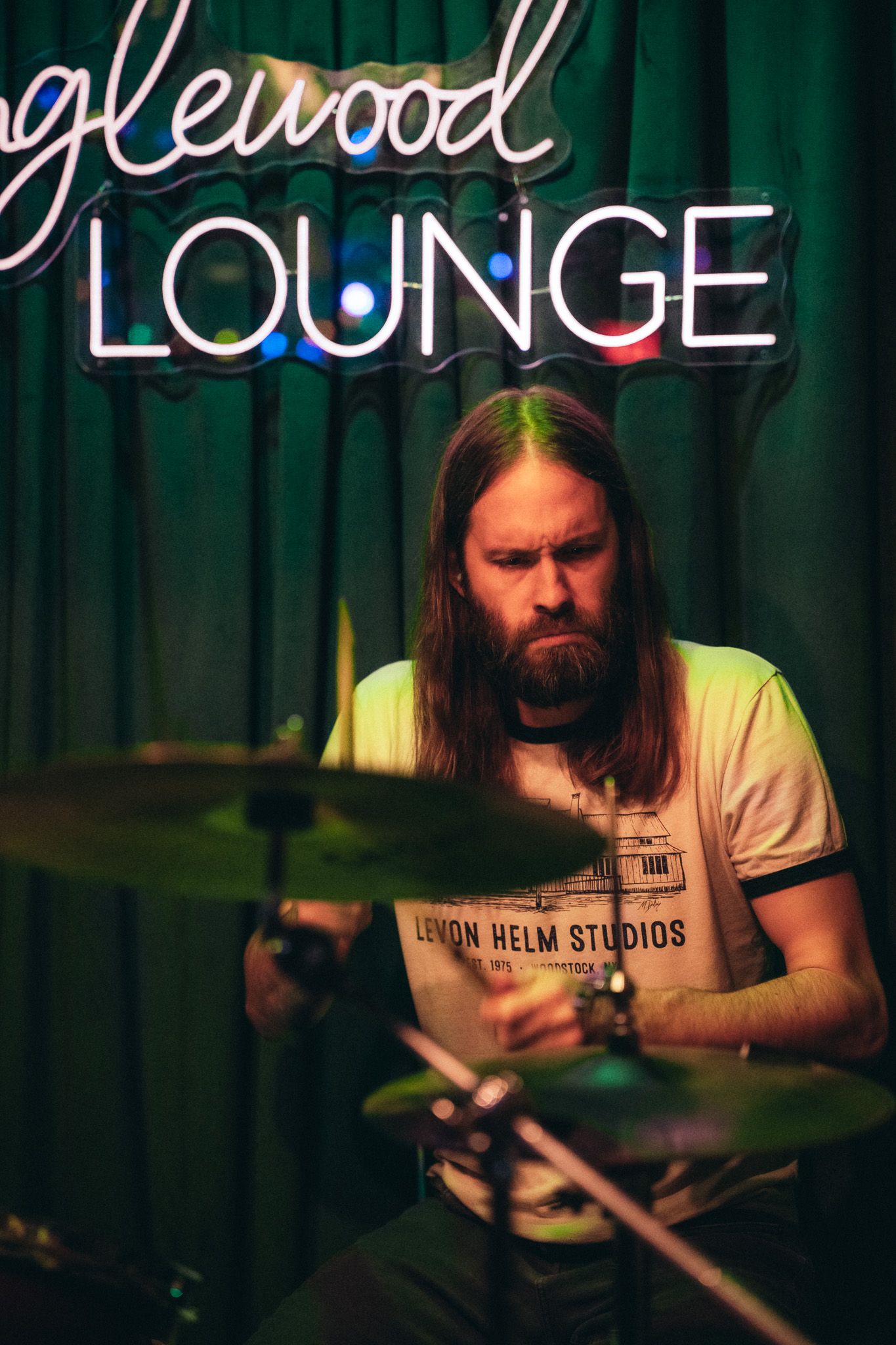 Bearded drummer with long hair playing drums beneath Englewood Lounge neon sign.