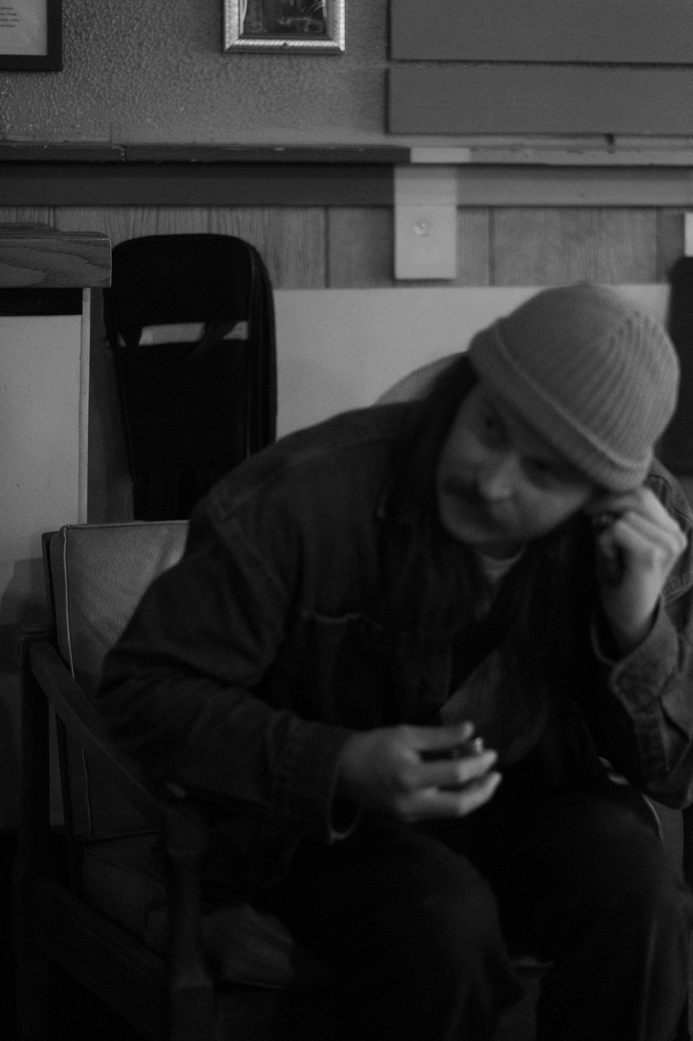 Man in beanie hat looks down at phone while seated in a chair in black and white photo.