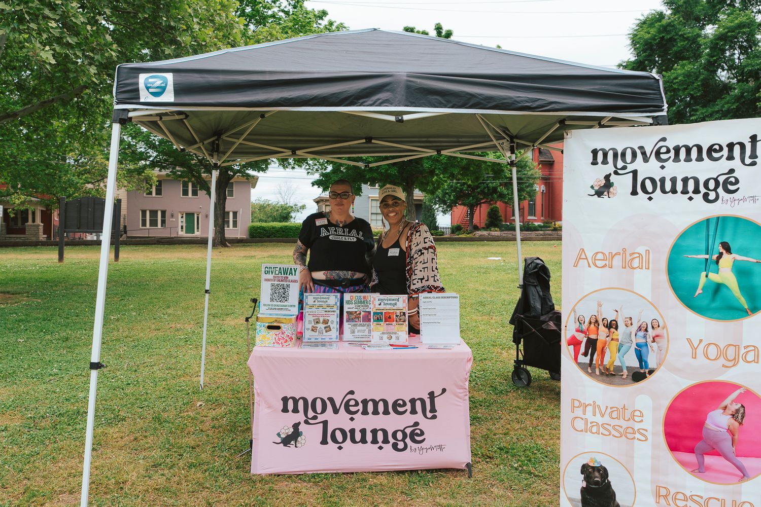 Two people at Movement Lounge outdoor booth promoting aerobics, yoga, and private fitness classes.