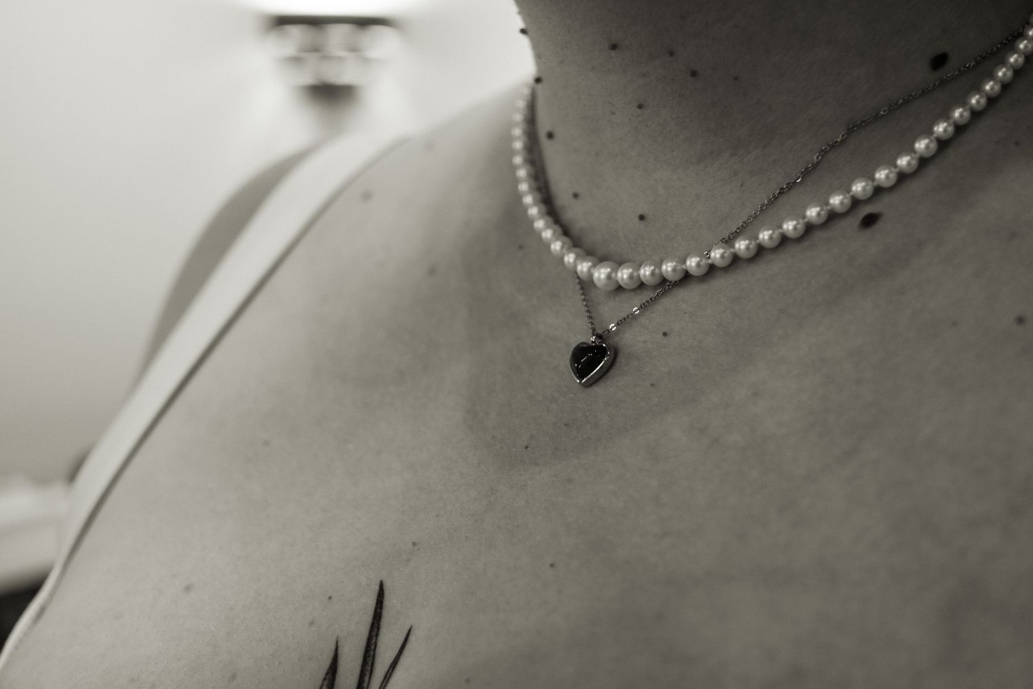 Black and white close-up of bride wearing pearl necklace with heart pendant and tattoo on chest.